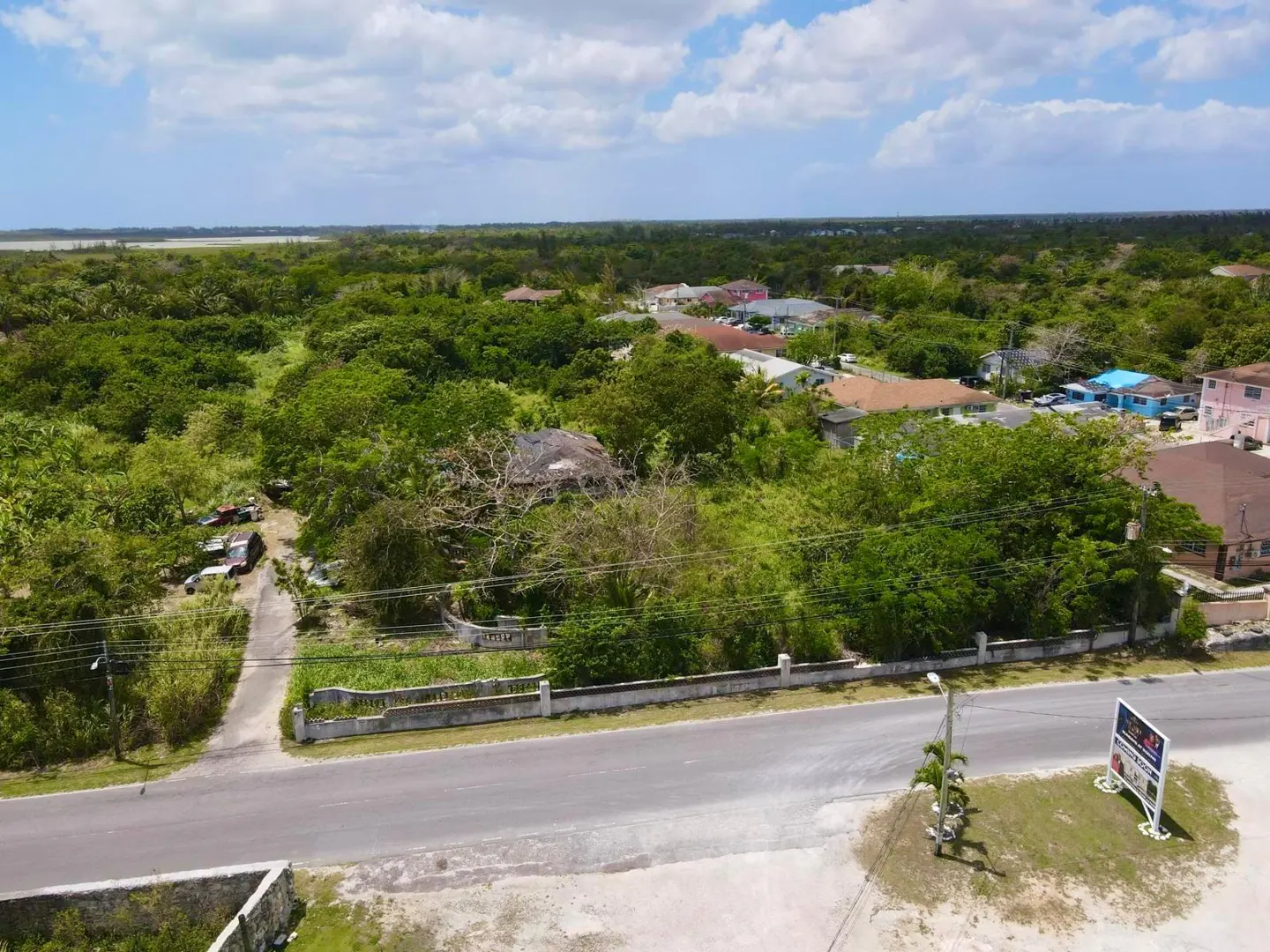 Aerial view of a lush, tree-filled residential lot next to a paved road with a real estate sign and distant houses.