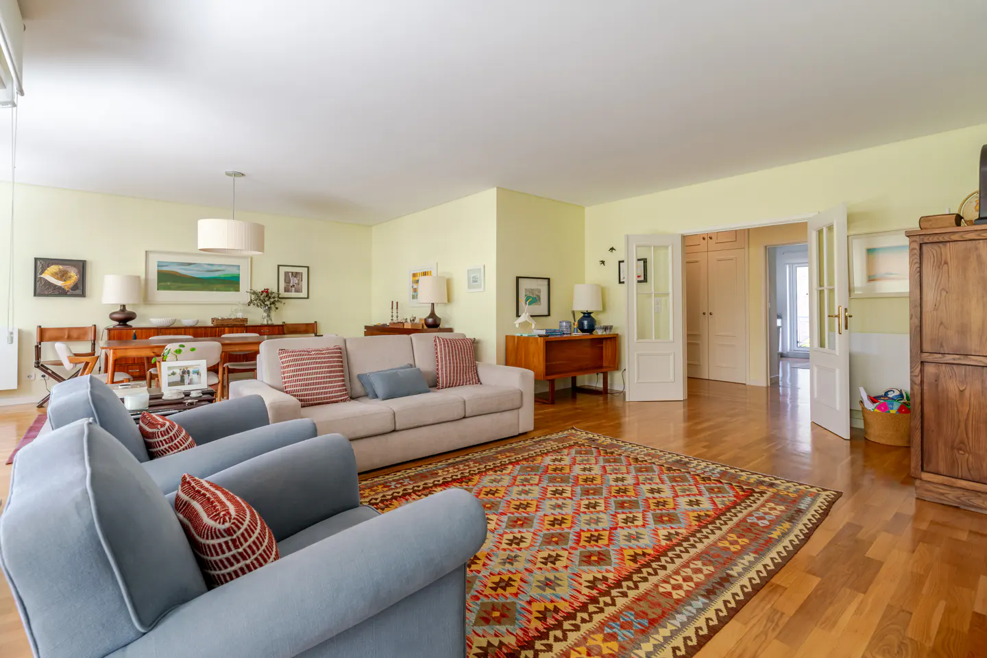Living room with light yellow walls, hardwood floors, and a colorful patterned rug. Blue armchairs and a beige sofa face each other. Dining area visible in the background.
