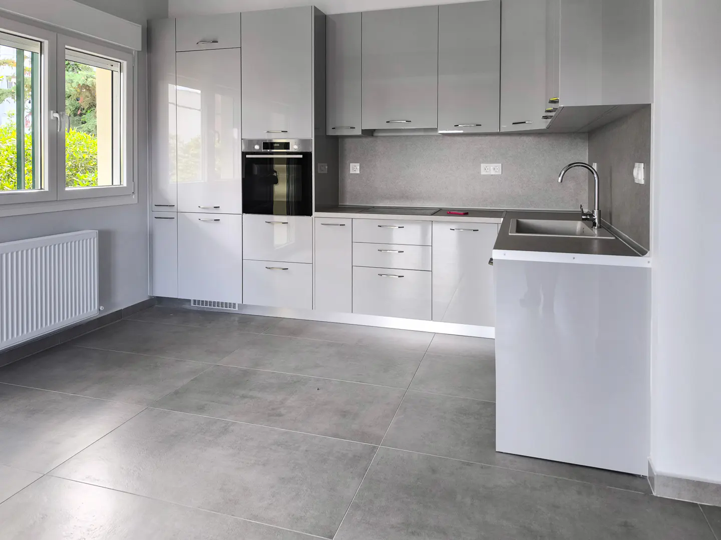 Modern kitchen with glossy light gray cabinets, stainless steel oven, sink, and large gray floor tiles near a window.