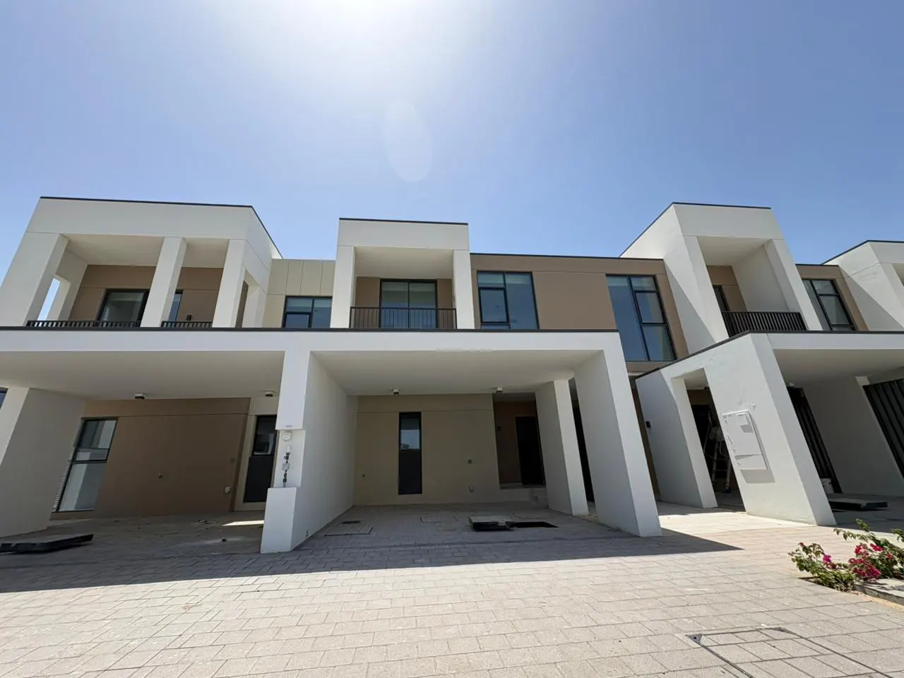 Exterior view of modern townhouses with white and beige facades, carports, and a paved driveway under a clear blue sky.