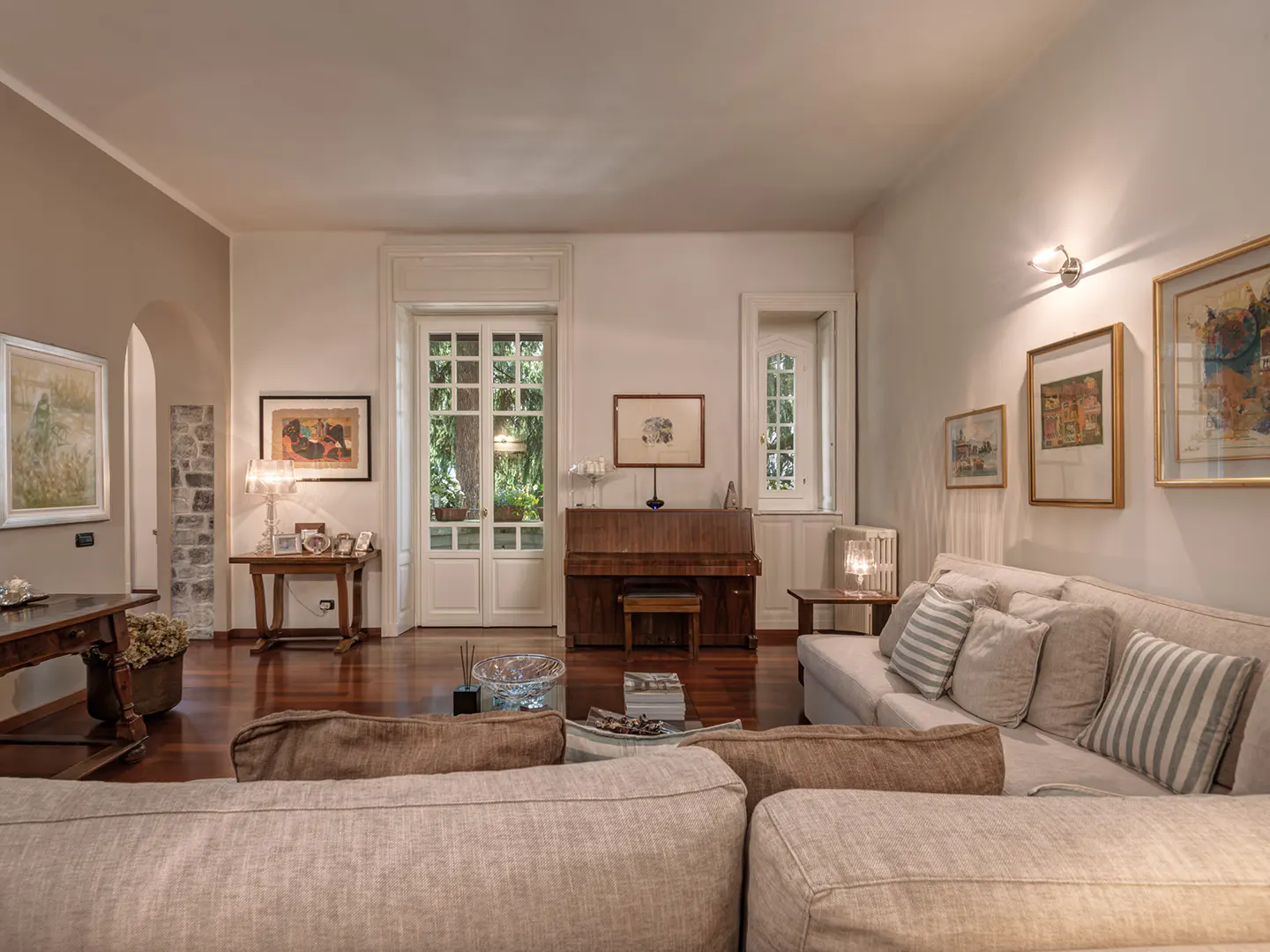 Elegant living room with hardwood floors, a beige sectional sofa, an upright piano, and framed art on white walls.