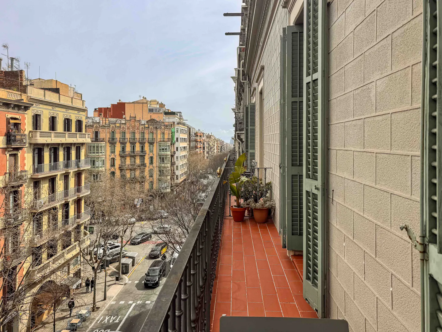 View from a balcony with terracotta tiles, green shutters, and potted plants overlooking a city street with cars and buildings.
