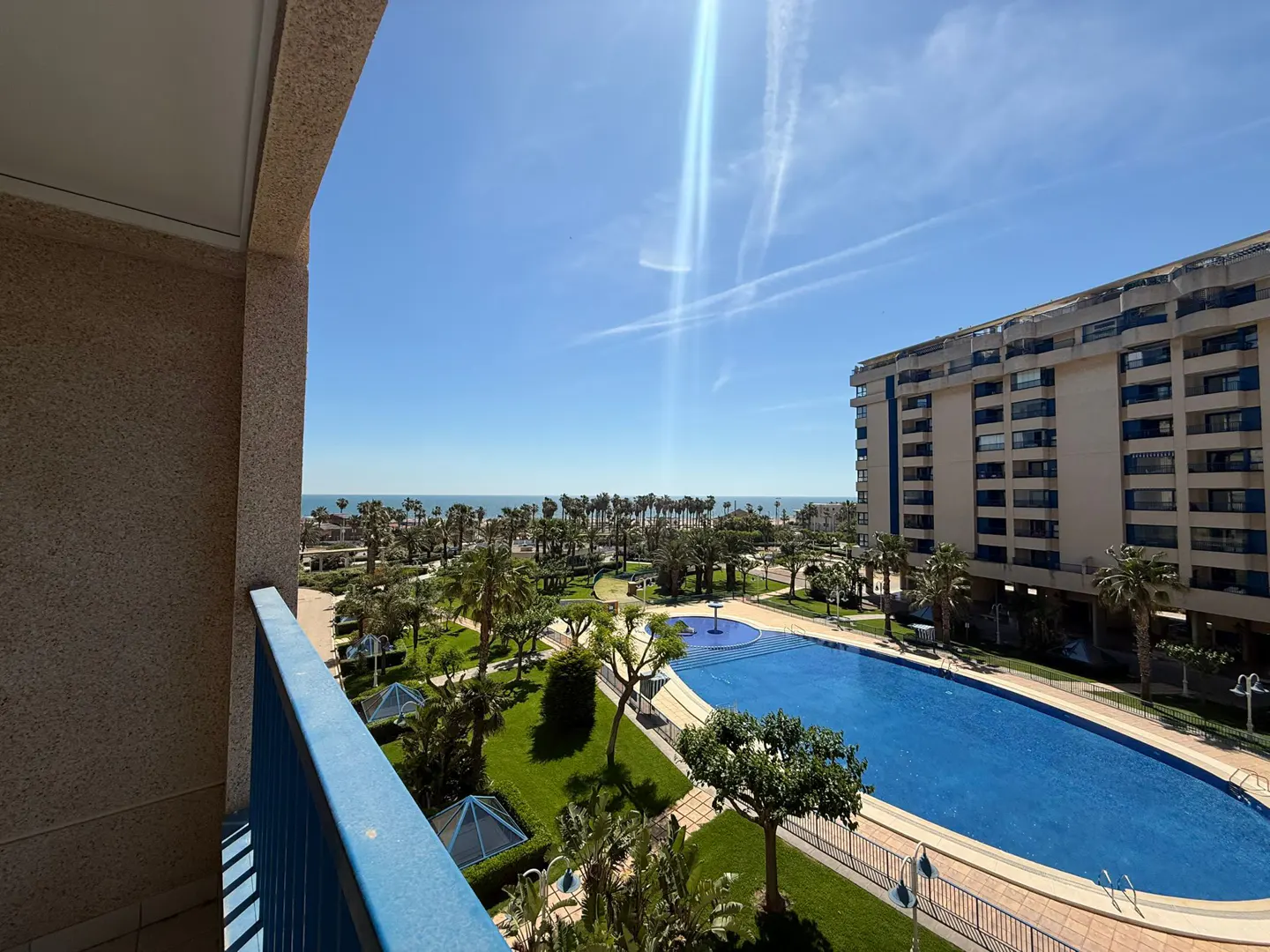 View from a balcony overlooking a pool, palm trees, and the ocean under a blue sky. A tall building is on the right.