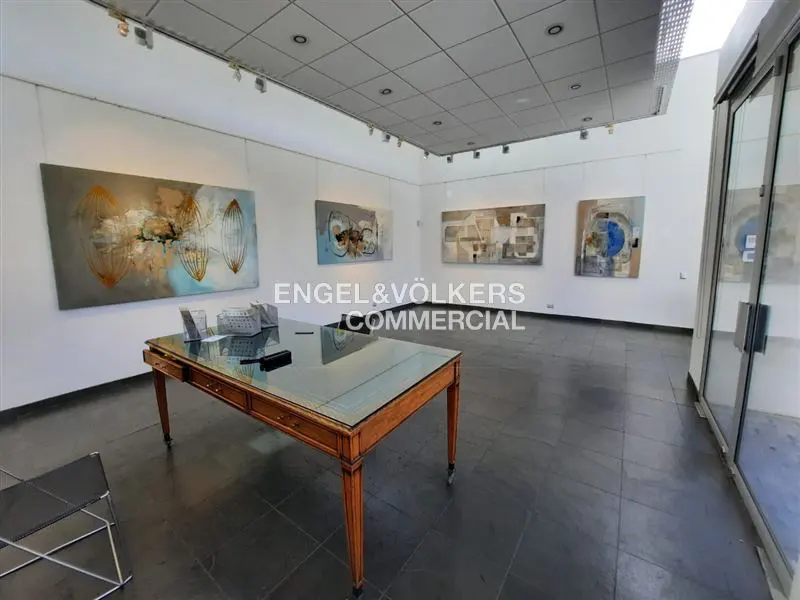 Gallery space with abstract art on white walls, dark floors, and a wooden table with a glass top. Natural light from a glass door.