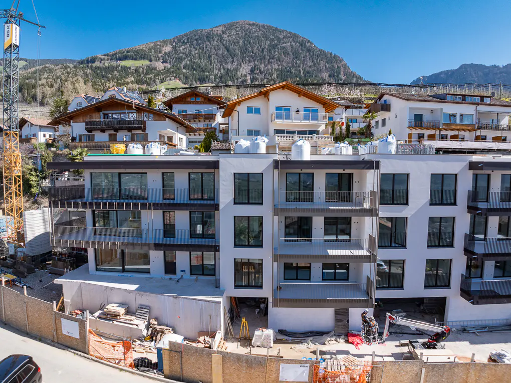 A modern, white apartment building under construction, with a mountain backdrop and blue sky. A yellow crane is visible on the left.