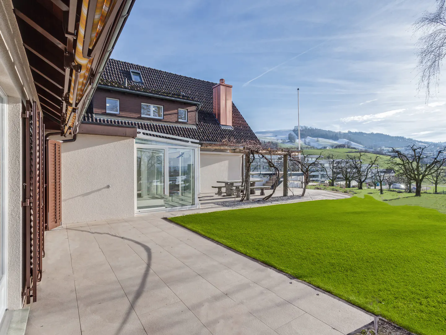 Exterior view of a house with a stone patio, green lawn, and a pergola with a stone table and benches. Rolling hills in the background.