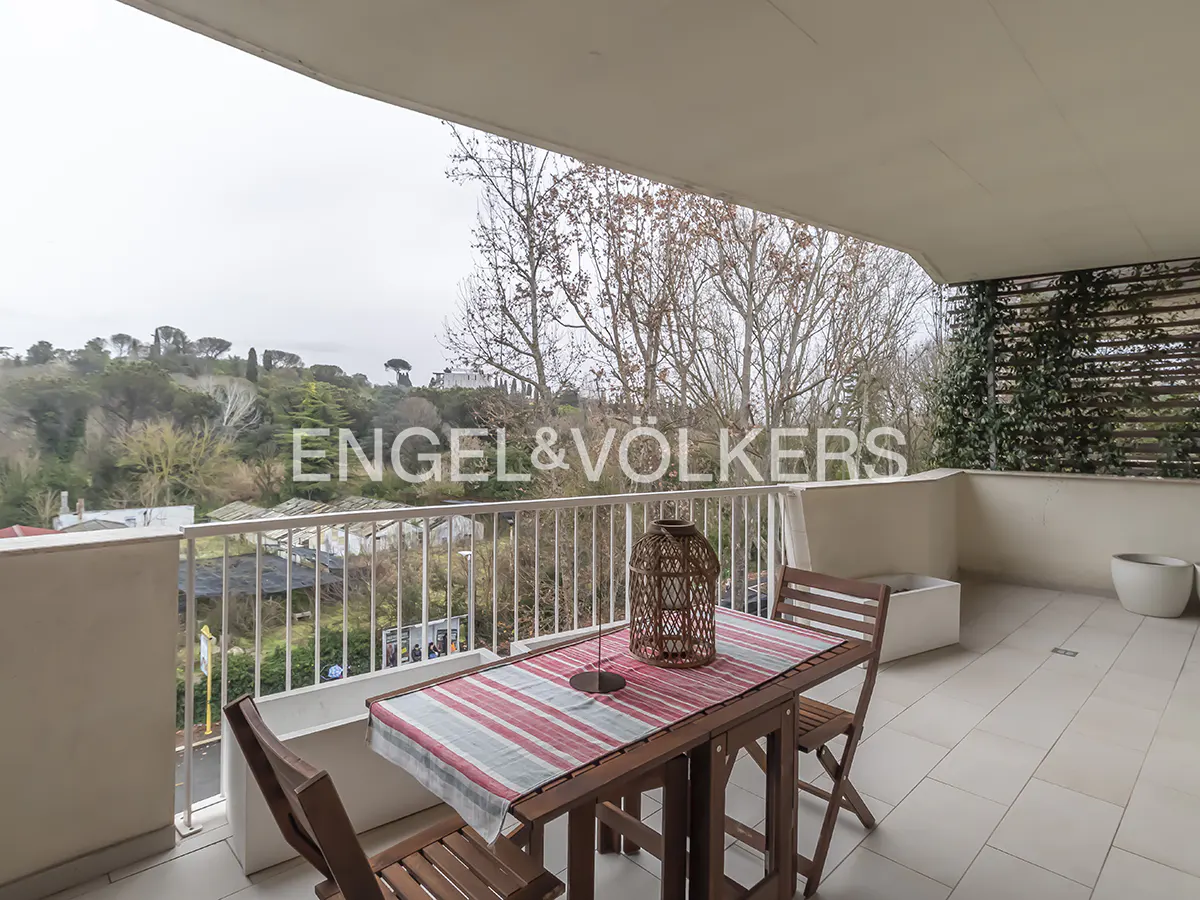 Balcony with a wooden table and chairs, a striped tablecloth, and a wicker lantern. A view of trees and a hill in the background.