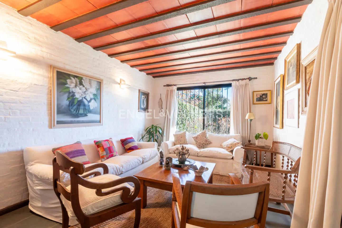 Cozy living room with white brick walls, terracotta beamed ceiling, two white sofas, wooden chairs, and a coffee table.