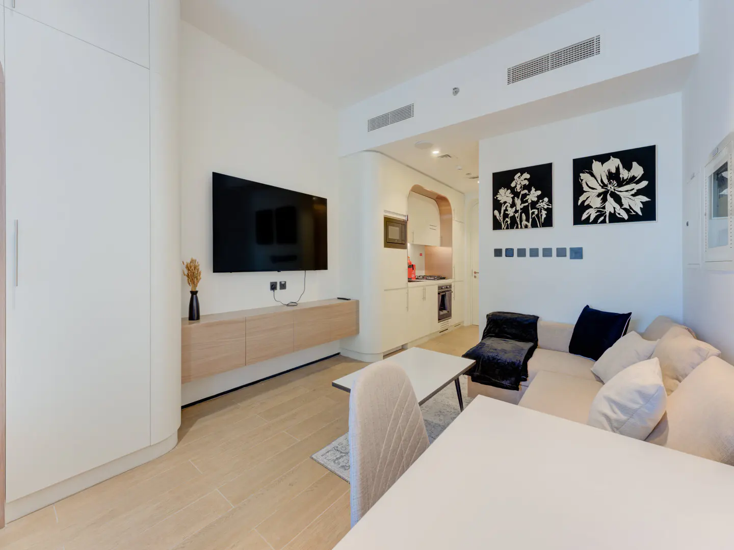 Bright apartment interior with a beige sofa, white table, TV, and kitchen. Black floral art hangs on the white wall.