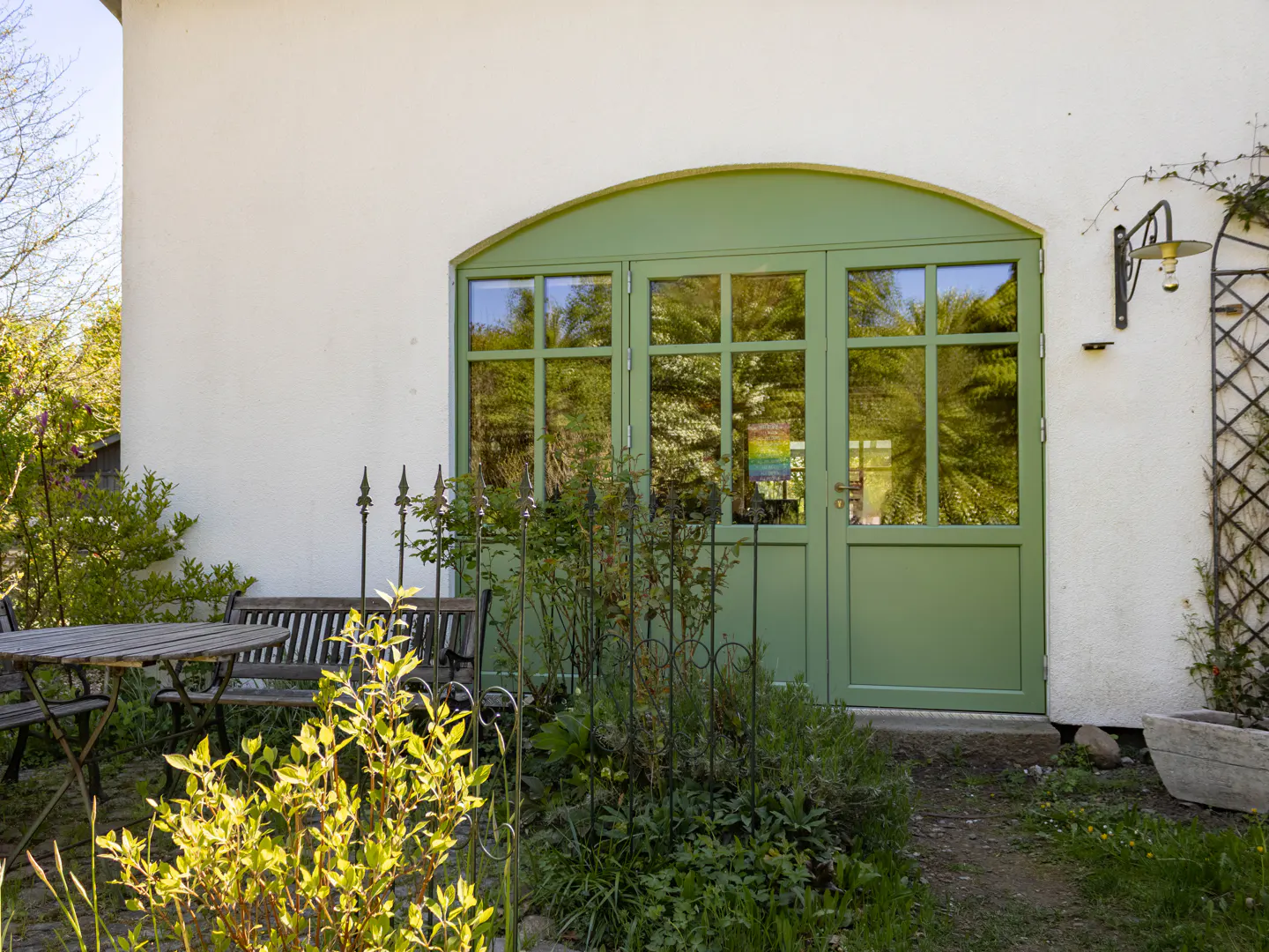 Exterior view of a white building with an arched green door and window. A table and bench sit on a stone patio surrounded by greenery.
