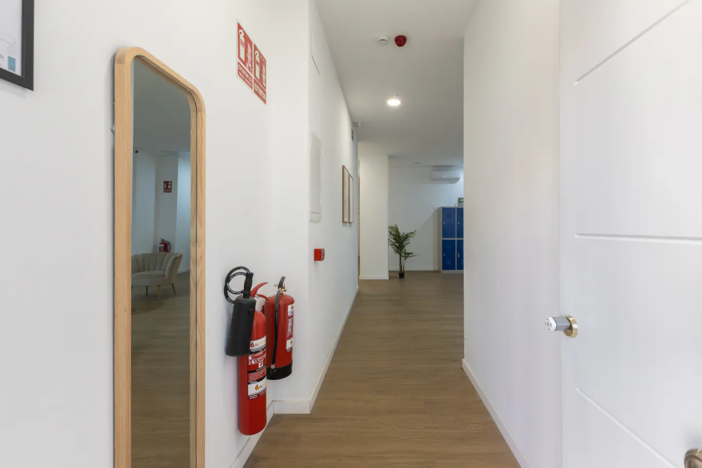 Hallway with white walls, wood floor, mirror, and fire extinguishers. Blue lockers are visible at the end of the hall.