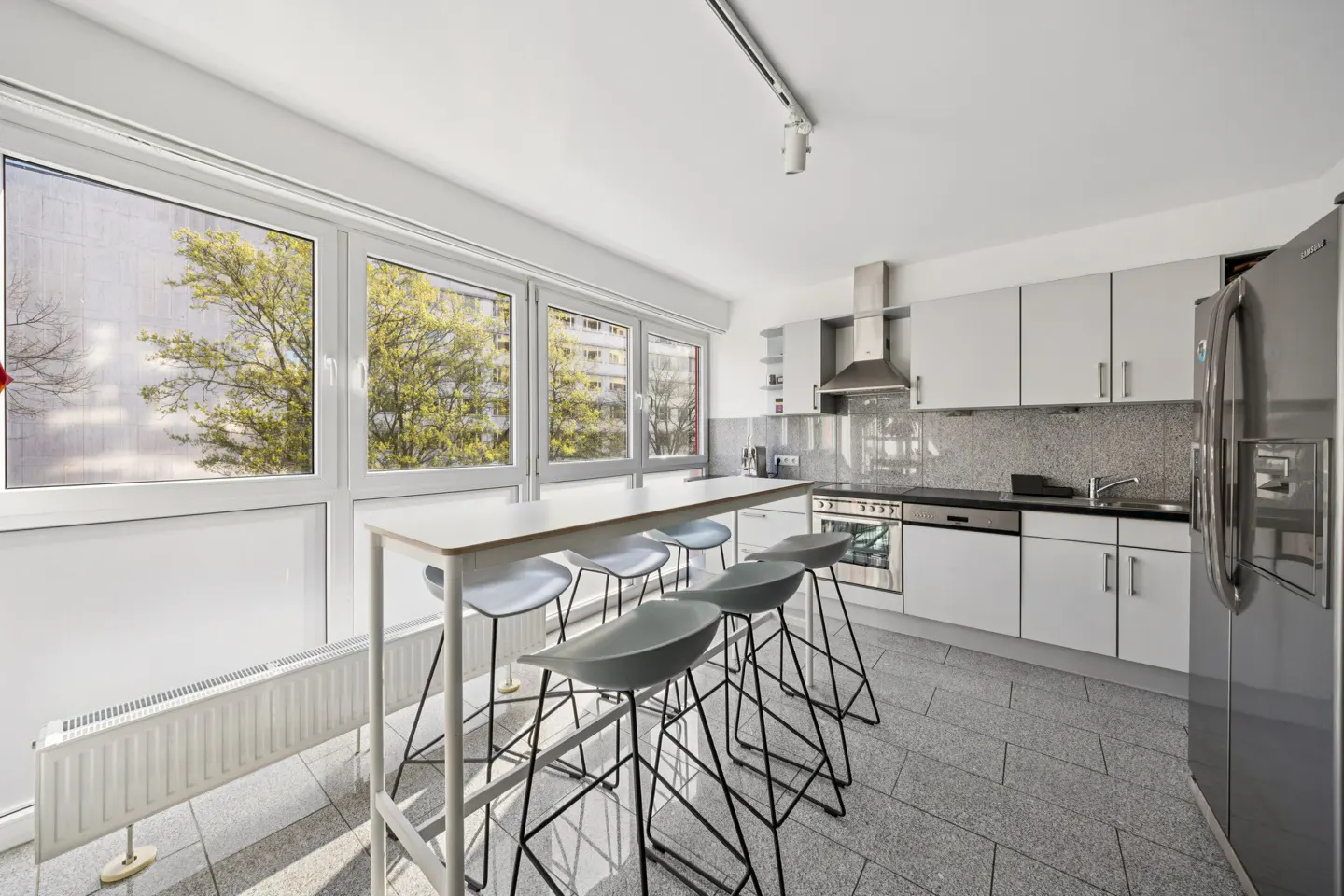 Bright kitchen with white cabinets, stainless steel appliances, and a long table with six gray stools by a large window.