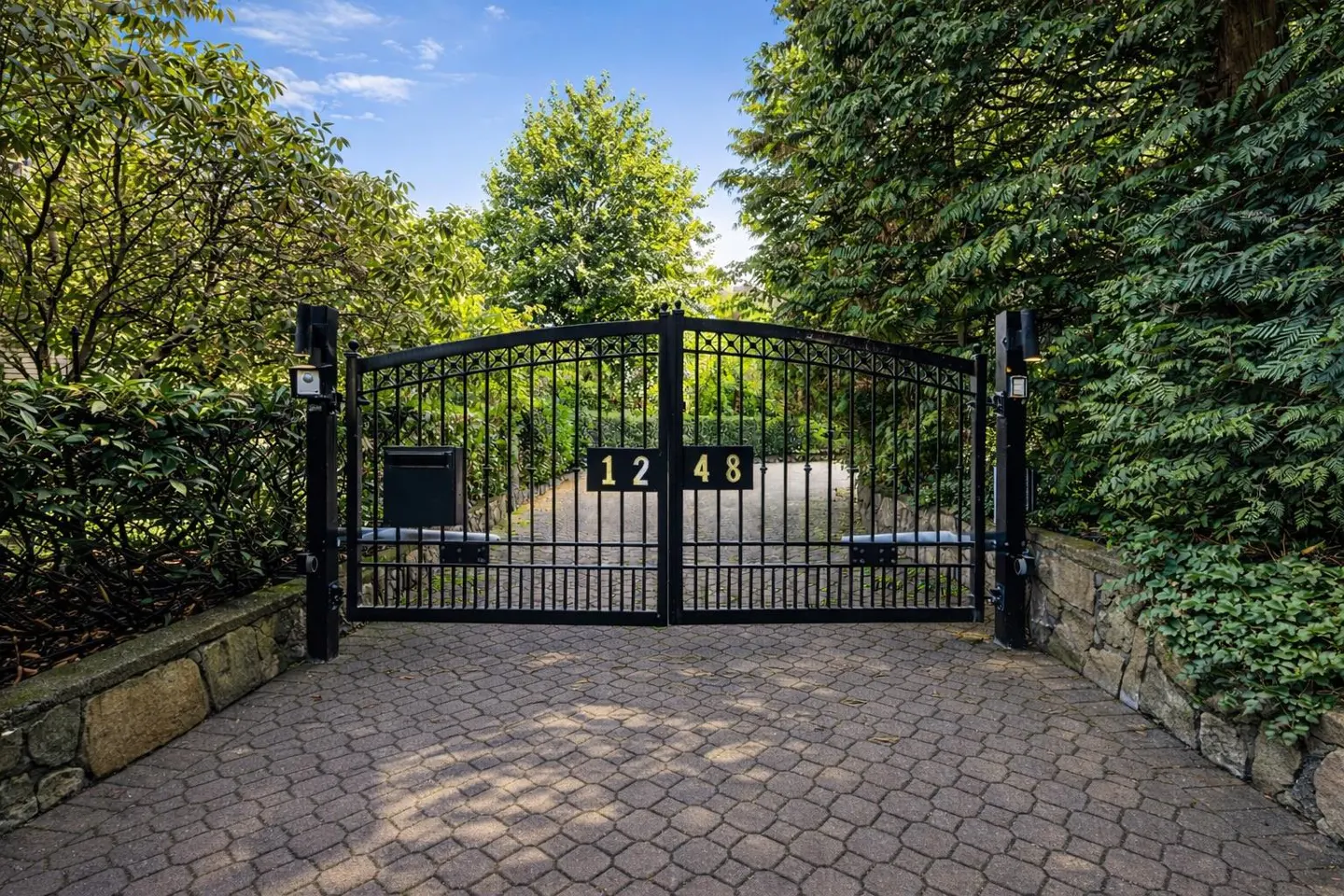 Black metal gate with the number 1248 in gold, set in a stone wall and surrounded by green trees. Paved driveway.