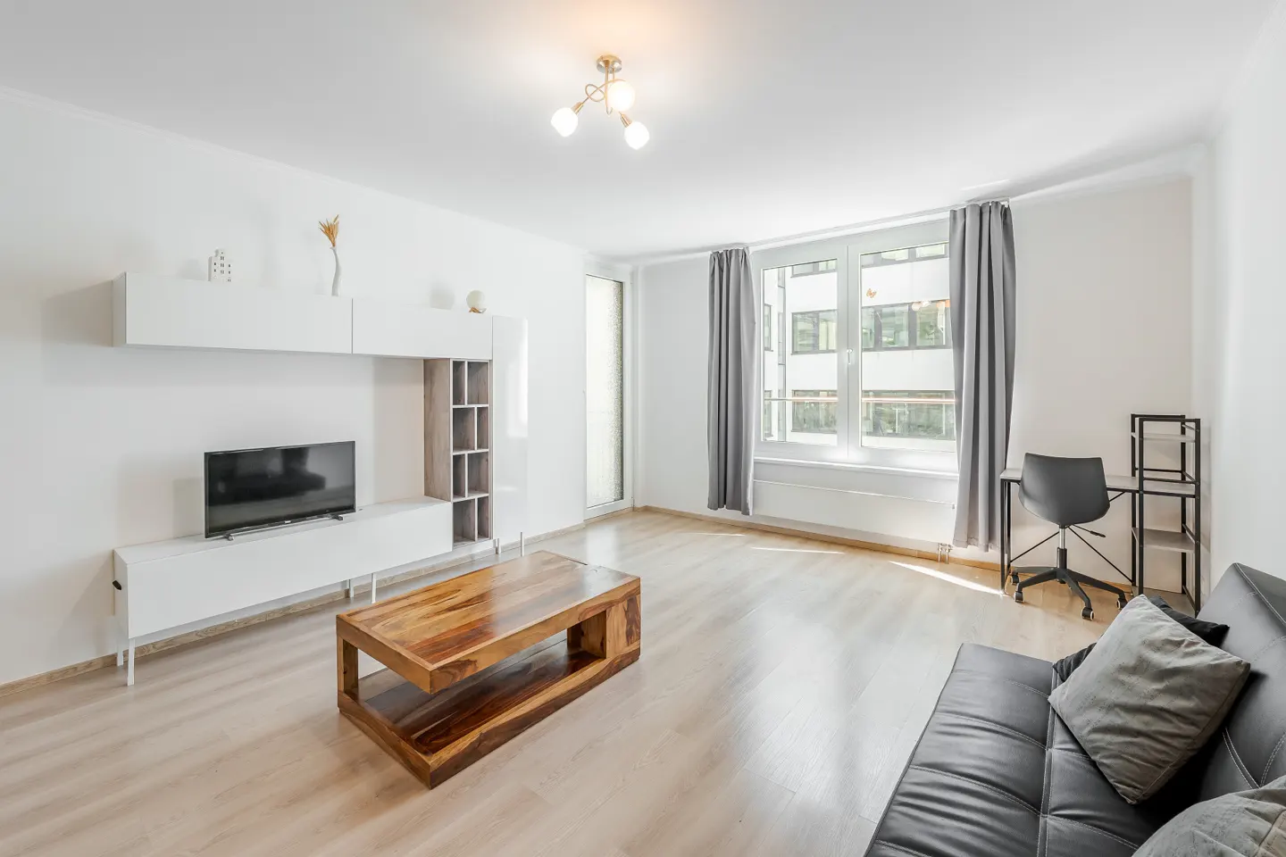 Bright living room with white walls, light wood floors, and a dark leather sofa. A wood coffee table sits in front of a white media center with a TV. A desk and chair are by the window.