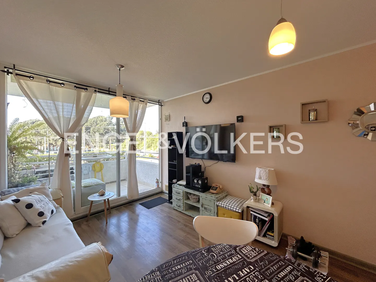 Living room with beige walls, wood floors, and a balcony. A sofa, TV, and side tables furnish the space. Natural light streams in through the balcony doors.