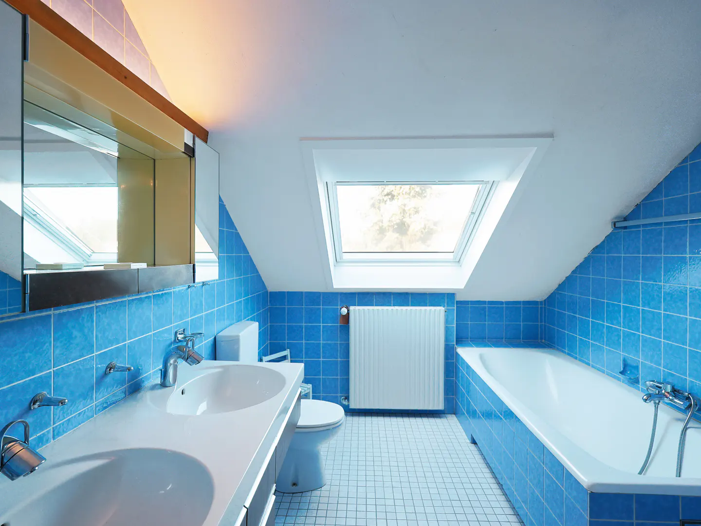 Bright blue tiled bathroom with double sink, toilet, bathtub, and skylight.