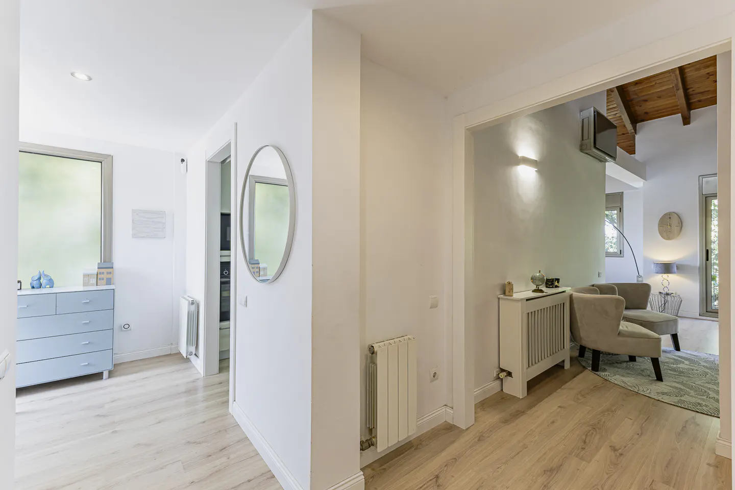 Bright, modern home interior with white walls and light wood floors. A blue dresser sits in a hallway, and a living room with armchairs is visible through an archway.