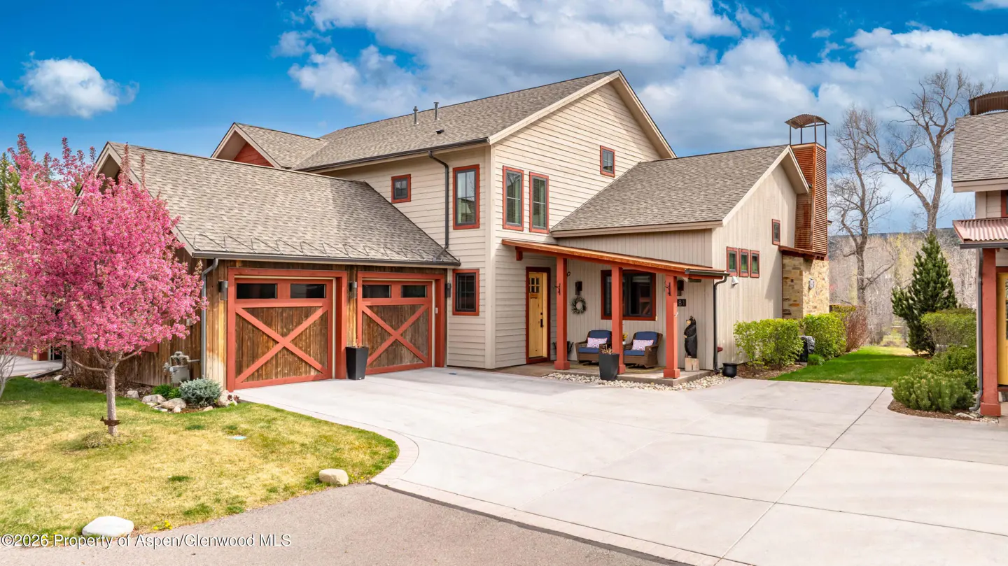 Exterior view of a two-story beige house with a brown roof, red trim, and a two-car garage with wooden doors. A pink tree is on the left.