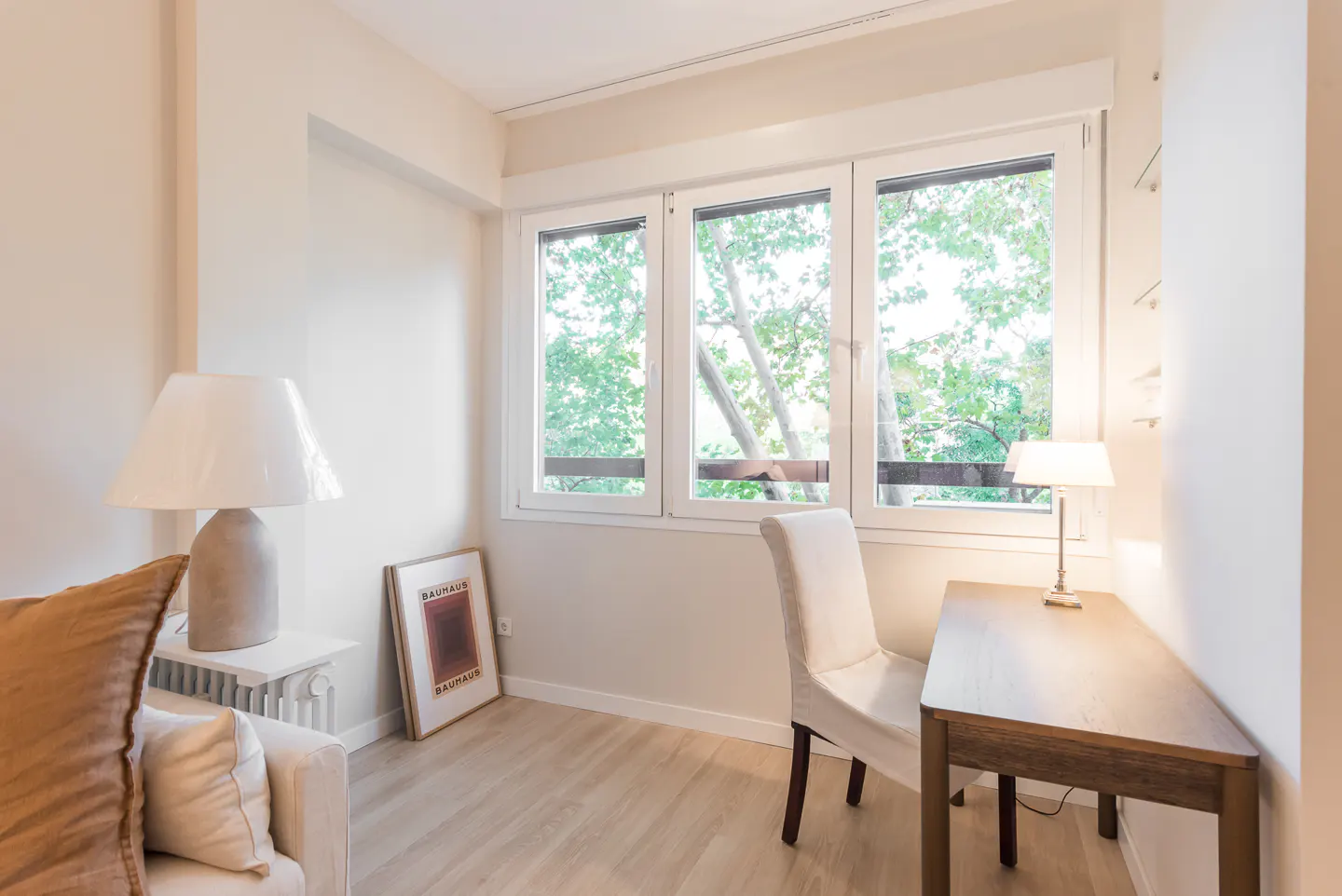 Bright room with a desk, chair, and large window showing green trees. A lamp sits on the desk, and another is on a side table next to a sofa. A framed Bauhaus poster leans against the wall.