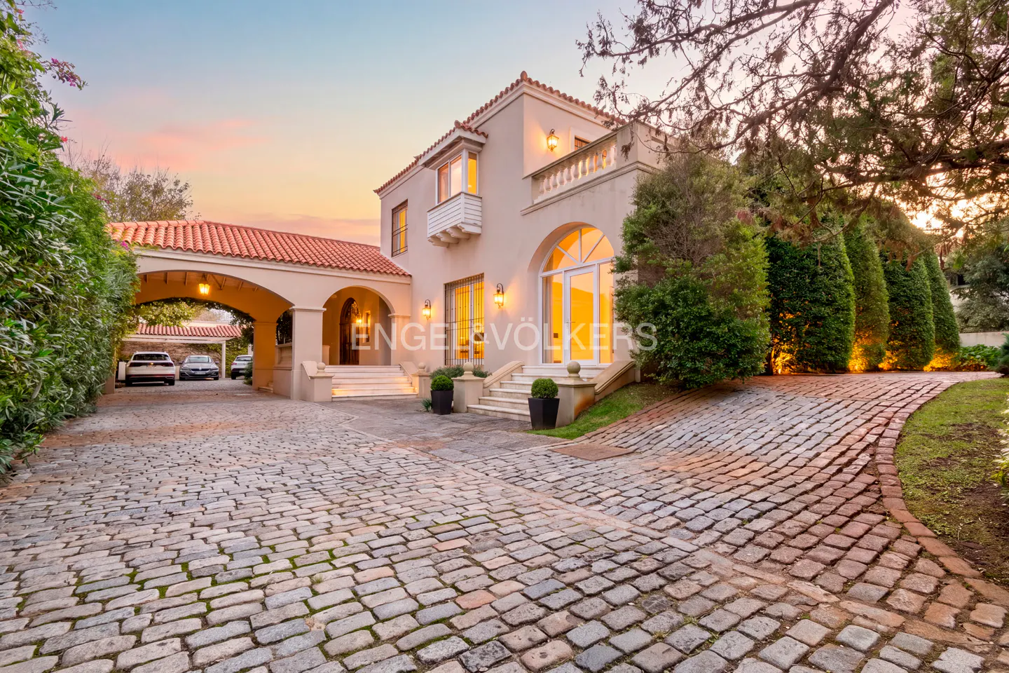 Exterior view of a two-story beige house with a red tile roof and a cobblestone driveway at sunset.