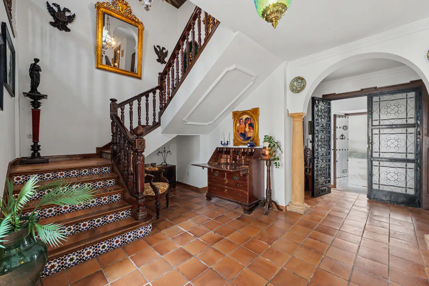 Ornate foyer with terracotta tile floor, wooden staircase with patterned tile risers, and antique desk. Arched doorway with iron gates.