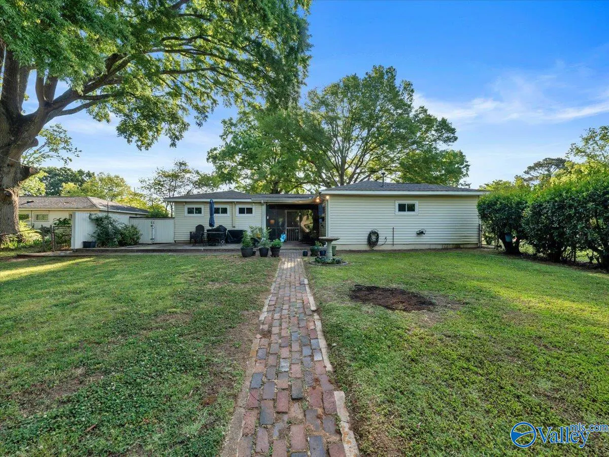 Backyard view of a one-story beige house with a brick path leading to a patio with furniture. Green grass and trees surround the house under a blue sky.