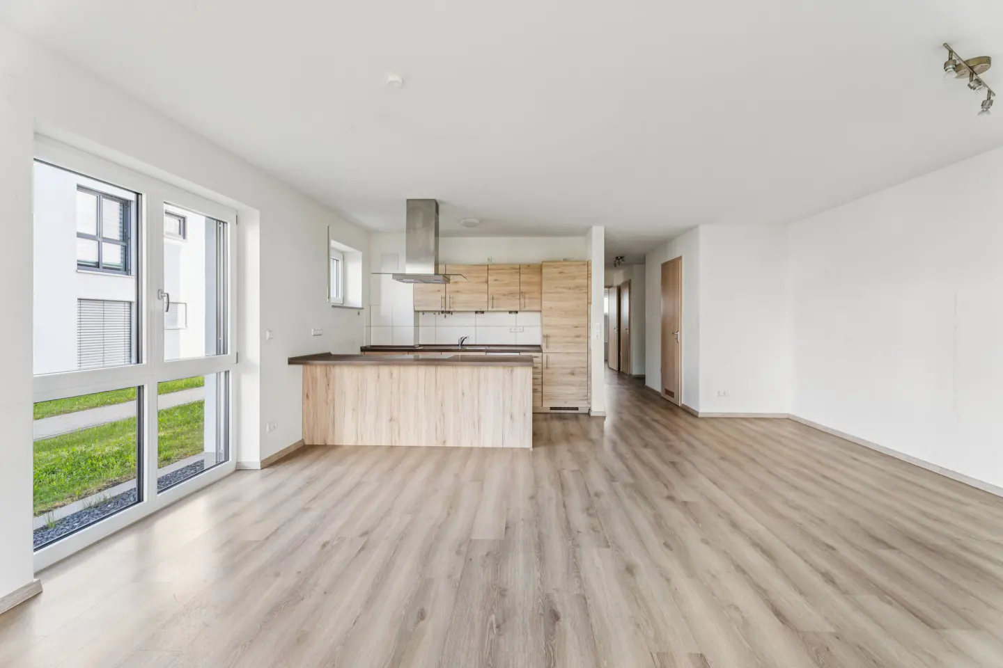 Bright, empty apartment with wood floors, white walls, and a kitchen with light wood cabinets. A large window shows a building and grass outside.