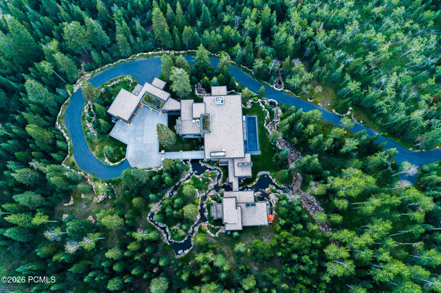 Aerial view of a modern home nestled in a dense green forest, featuring a winding driveway and a landscaped water feature.