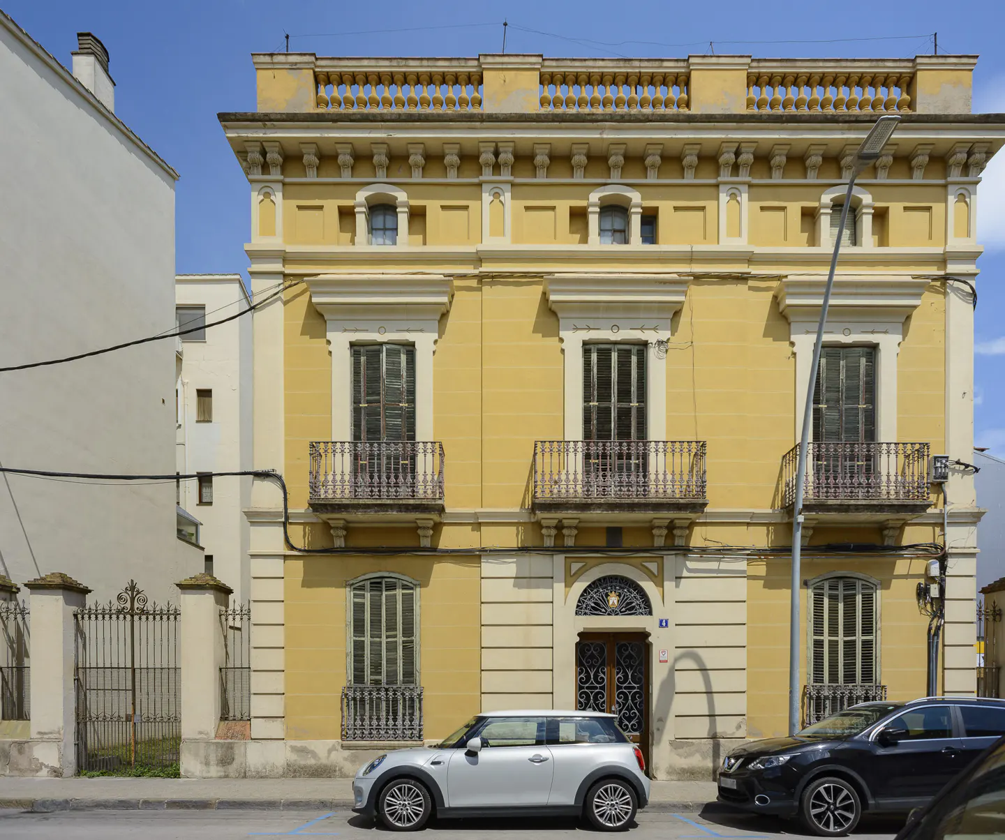 A three-story yellow historic building with ornate balconies and arched windows, featuring cars parked in front.