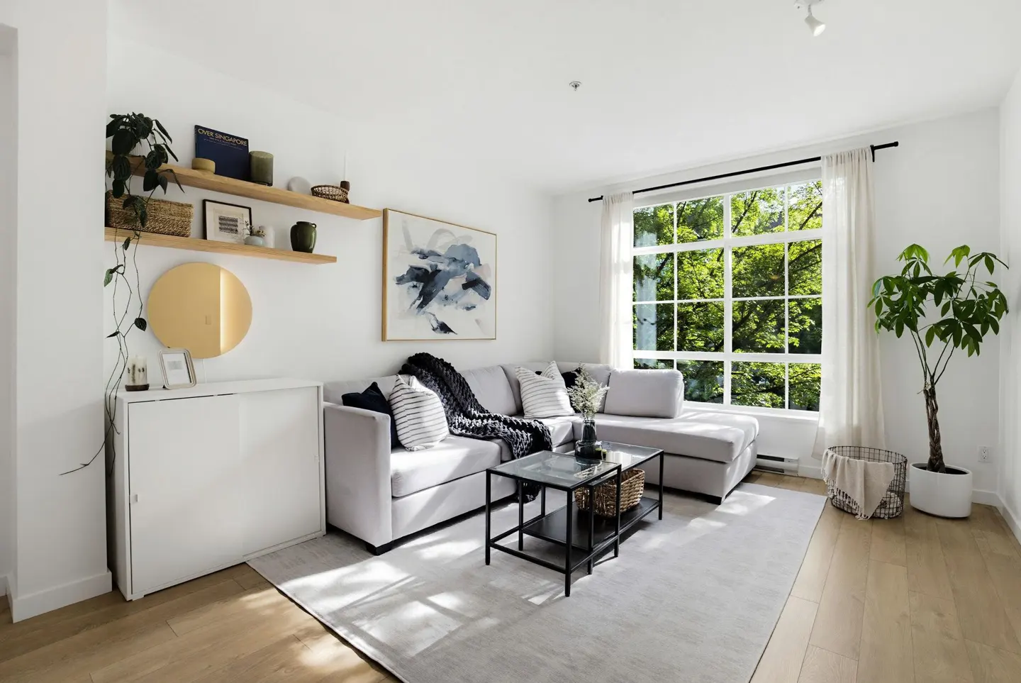 Bright living room with a gray sectional sofa, black coffee table, and large window with green trees outside. Shelves and art decorate the white walls.