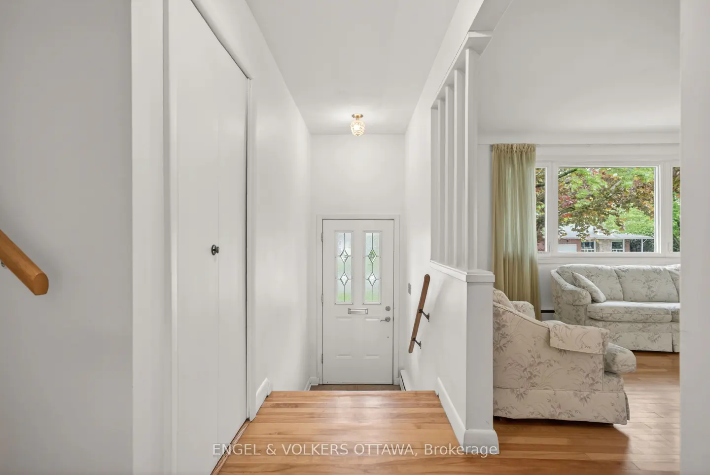 Interior view of a bright foyer with white walls, a closet, and a front door with glass panels. A living room with floral sofas is visible.