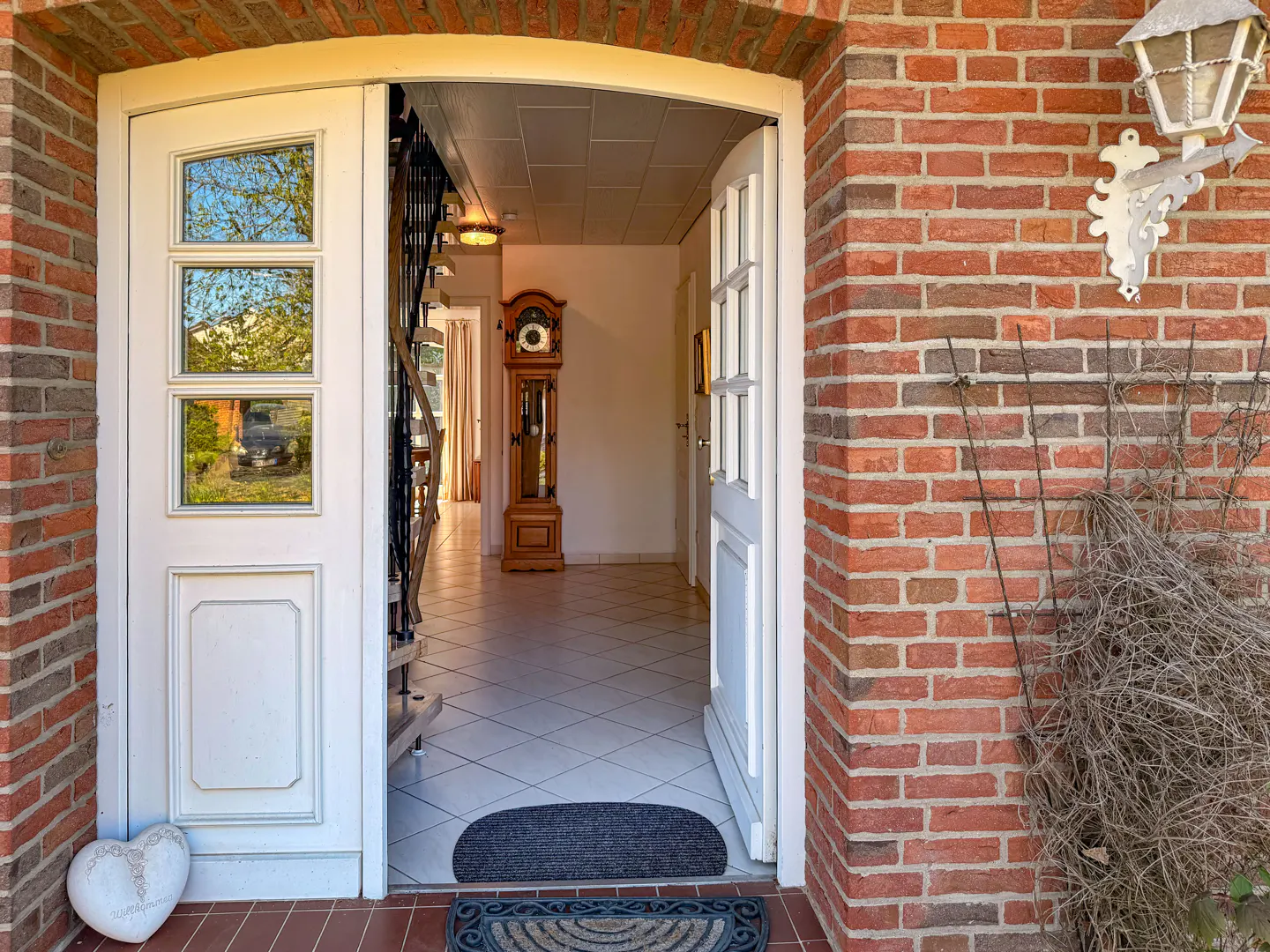 Open white front door of a brick house, revealing a tiled hallway with a grandfather clock and staircase.