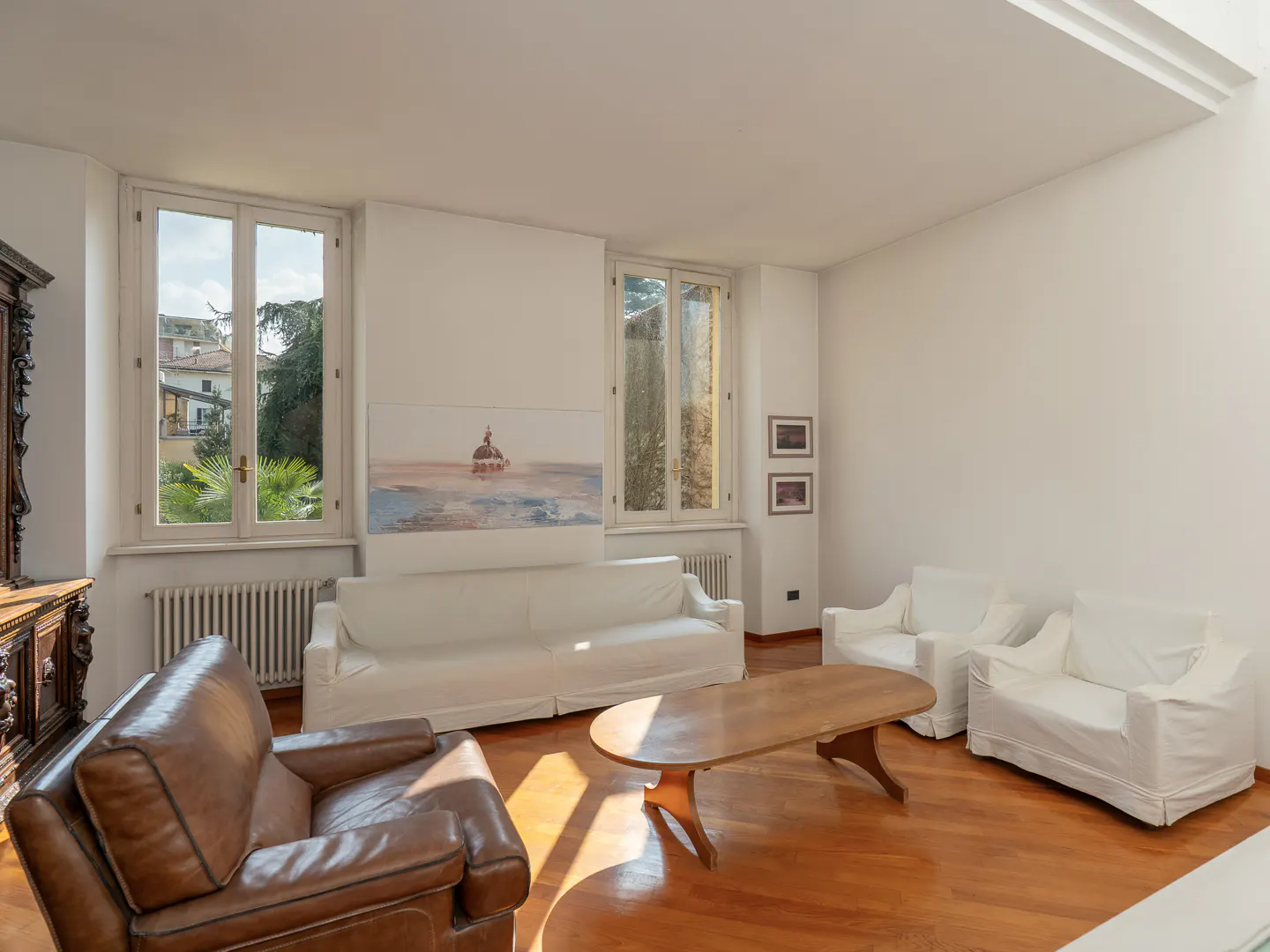 Bright living room with white sofas, a brown leather armchair, a wooden coffee table, and large windows on hardwood floors.
