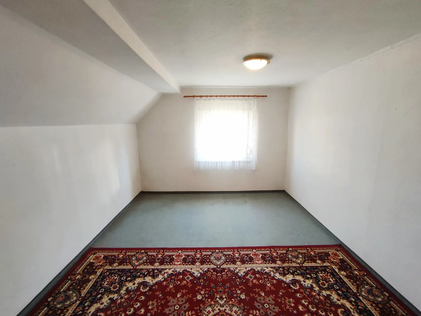 Attic room with white walls, a window with sheer curtains, and a red patterned rug on a green floor. A ceiling light is centered.