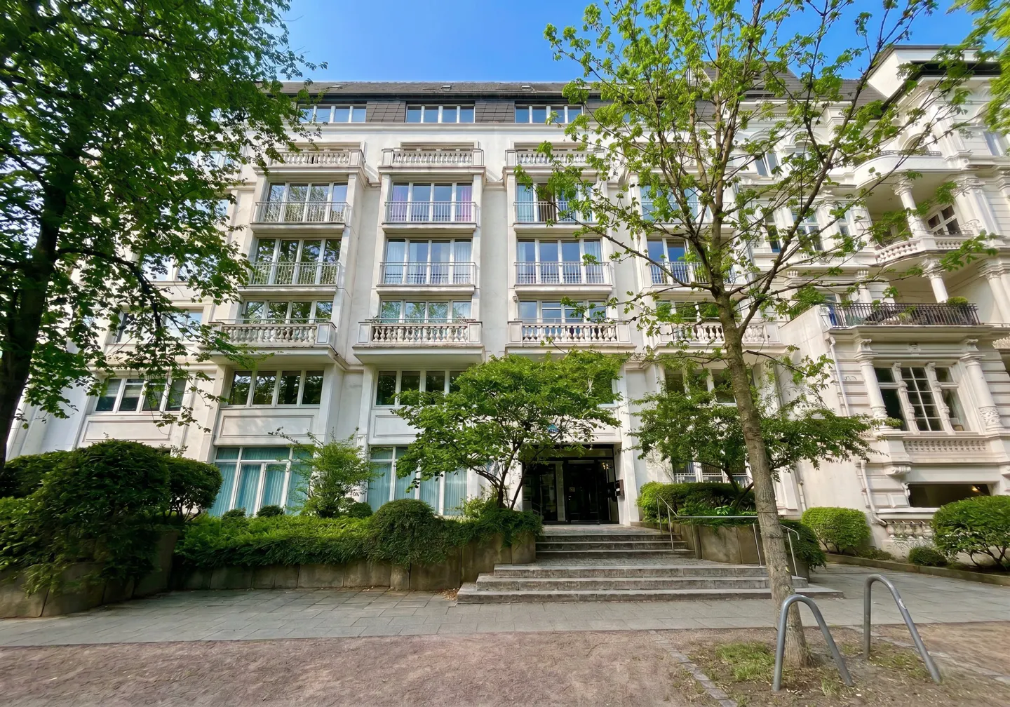 Exterior of a white apartment building with balconies, green trees, and a stone staircase leading to the entrance.