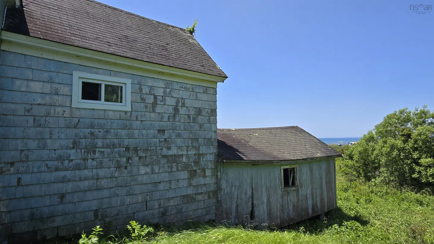 Side view of a weathered grey shingled house with a small attached shed, set in a grassy field overlooking the ocean.