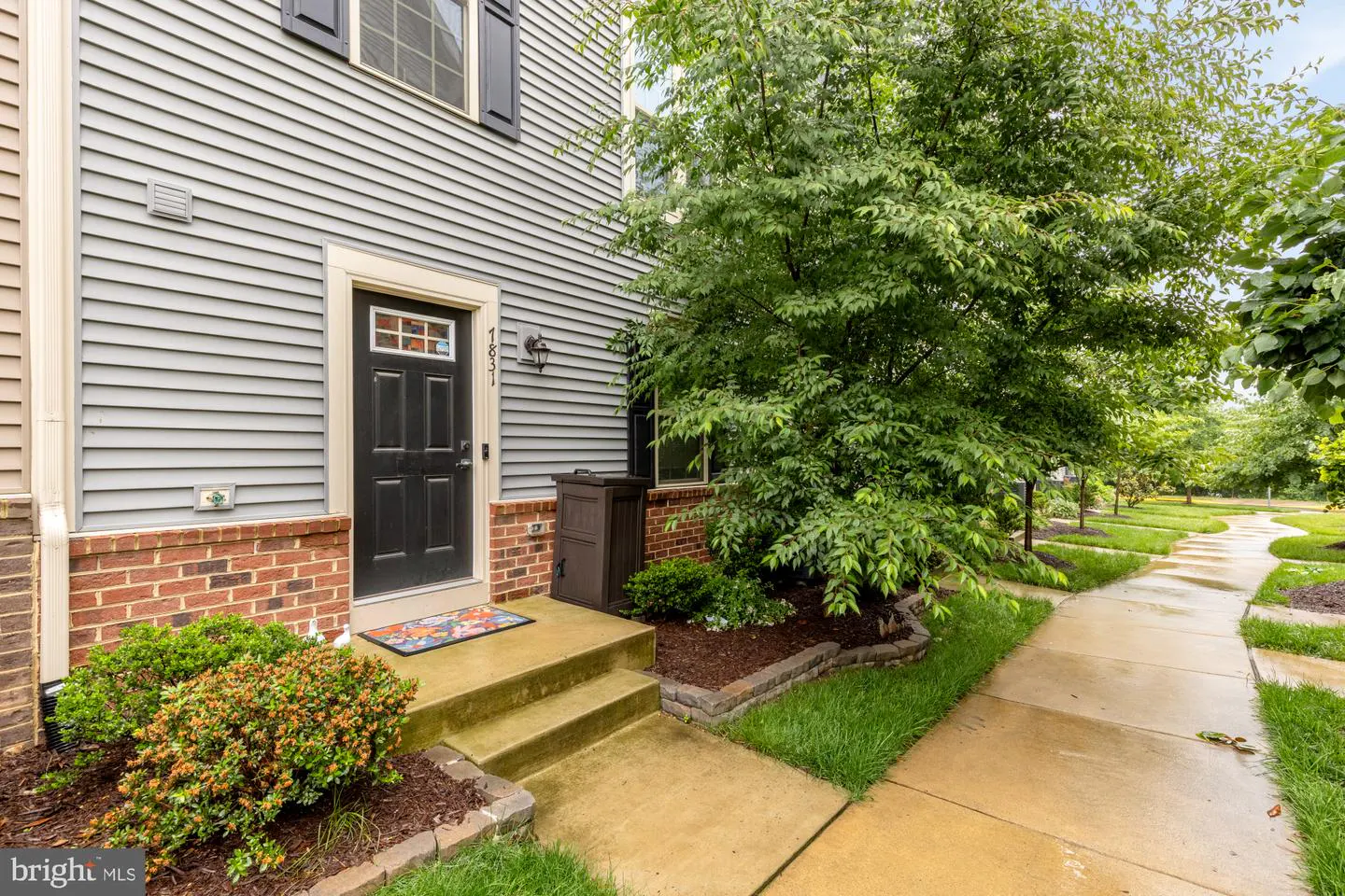 Exterior view of a gray house with a black door, brick foundation, and green landscaping.