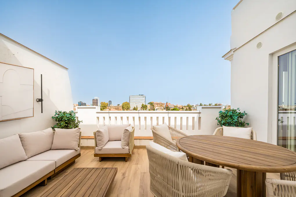 A bright, modern rooftop terrace with white walls, wooden furniture, and a city view.