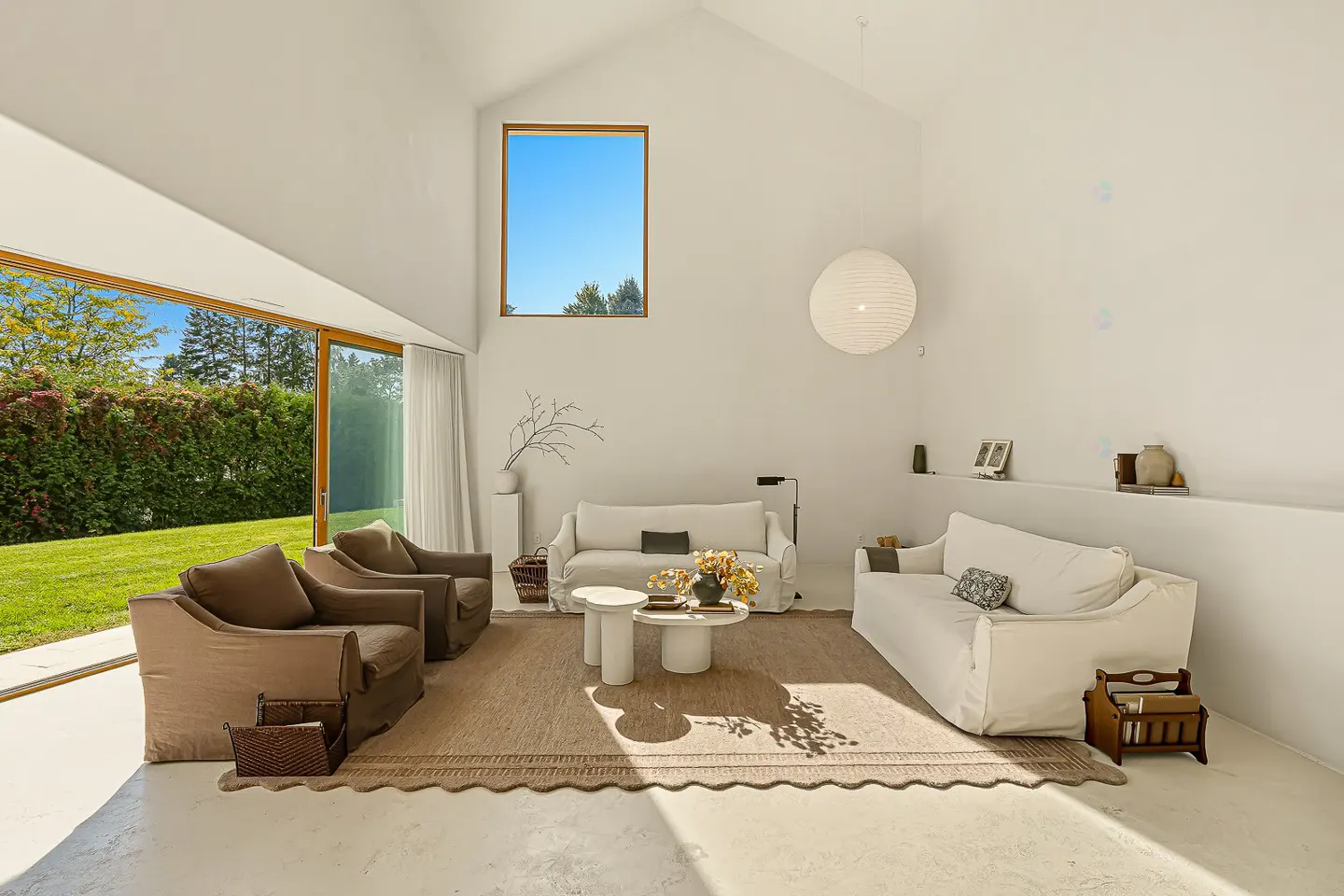 Bright, airy living room with white walls, brown armchairs, and white sofas. Large windows offer views of a green lawn and trees. A round paper lantern hangs from the ceiling.