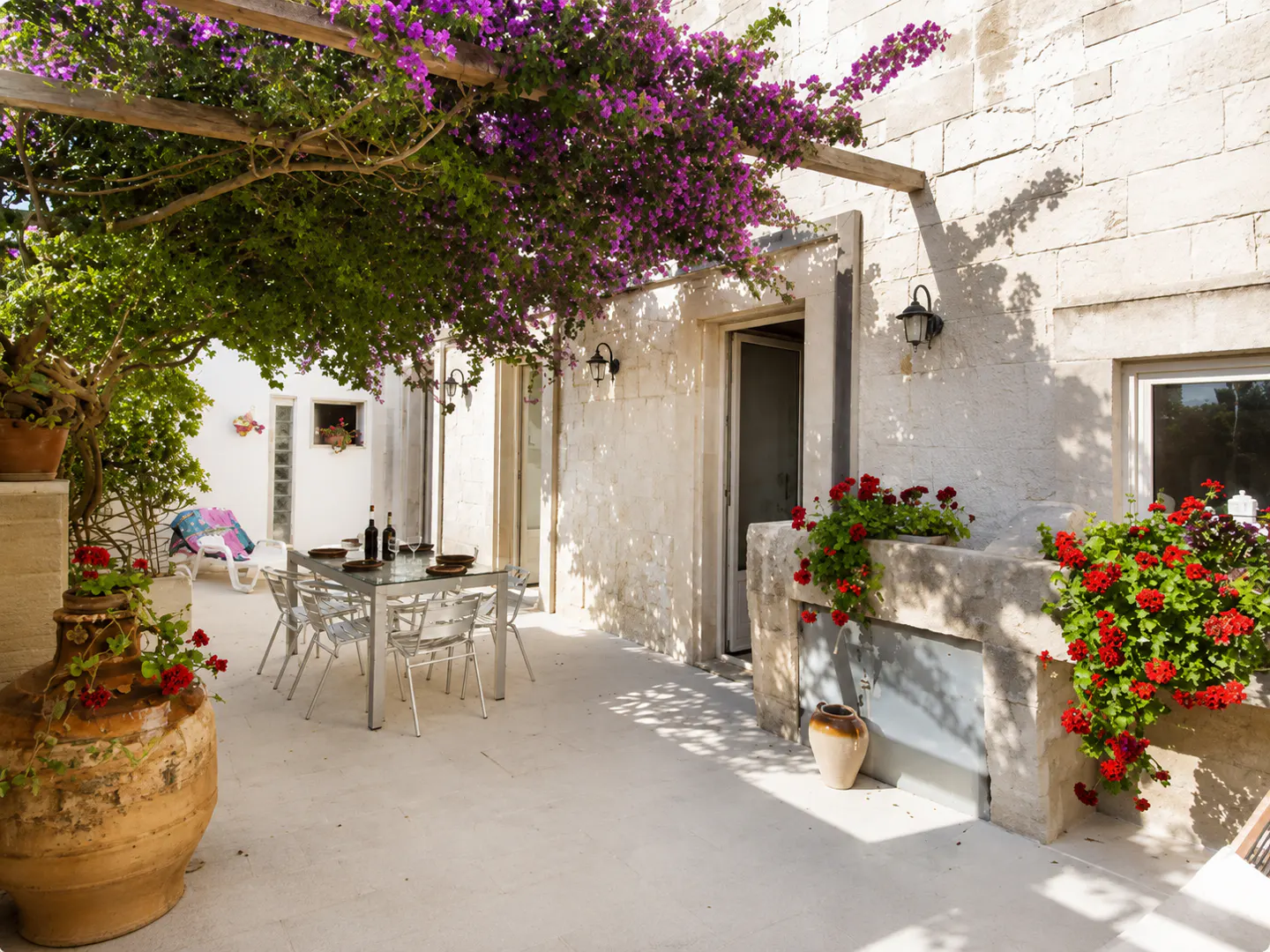 Outdoor patio with stone walls, purple bougainvillea, and a glass table set for dining. Red geraniums add color.