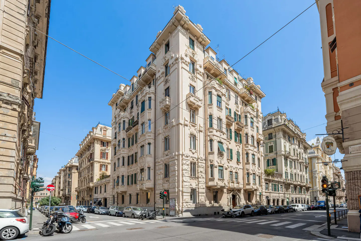 Street view of ornate, light-colored apartment buildings under a blue sky. Cars and motorcycles are parked along the street.