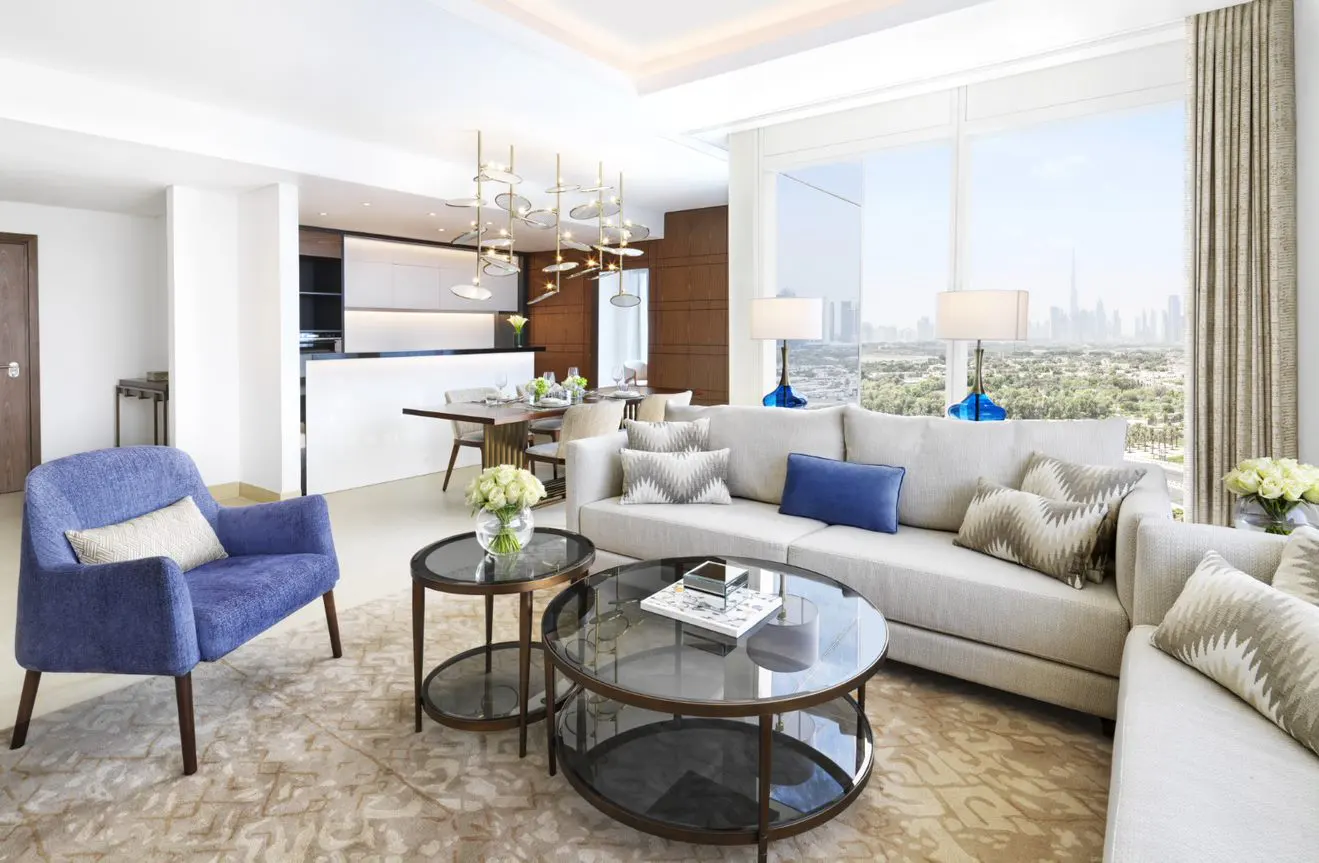 Modern living room with a beige sectional, blue armchair, glass coffee tables, and a view of the Dubai skyline.