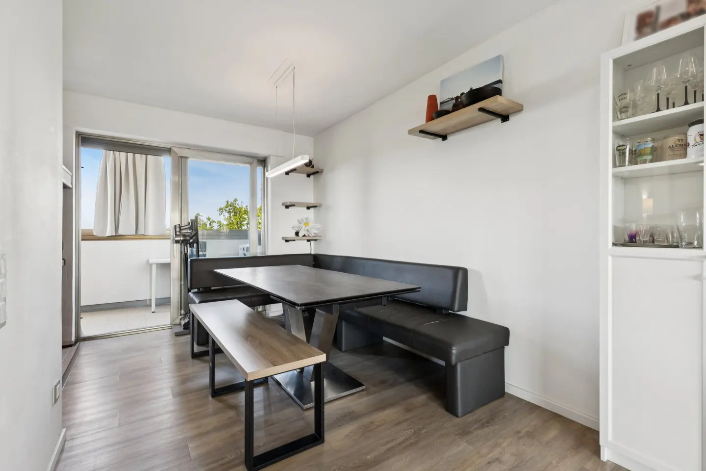 Bright dining area with a black table, bench seating, and a wooden bench. Shelves adorn the white wall, and a glass cabinet displays glassware.