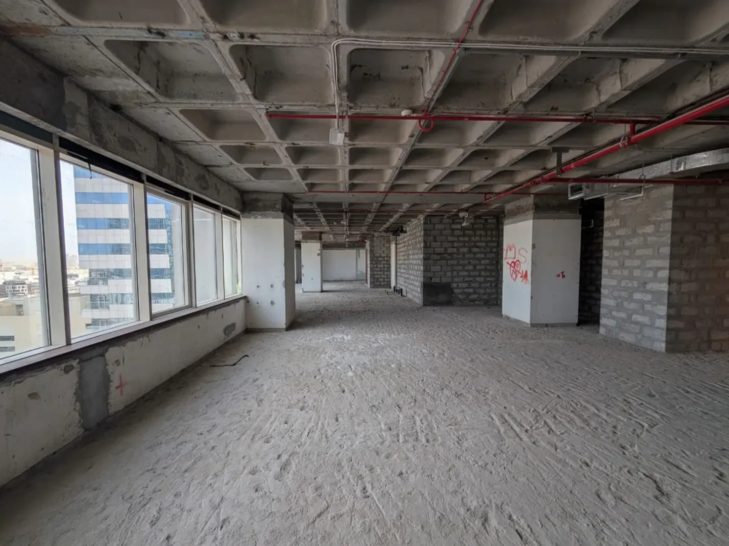 Unfinished office space with concrete floors, exposed ceiling, and brick walls. Large windows offer a city view. Red pipes run along the ceiling.