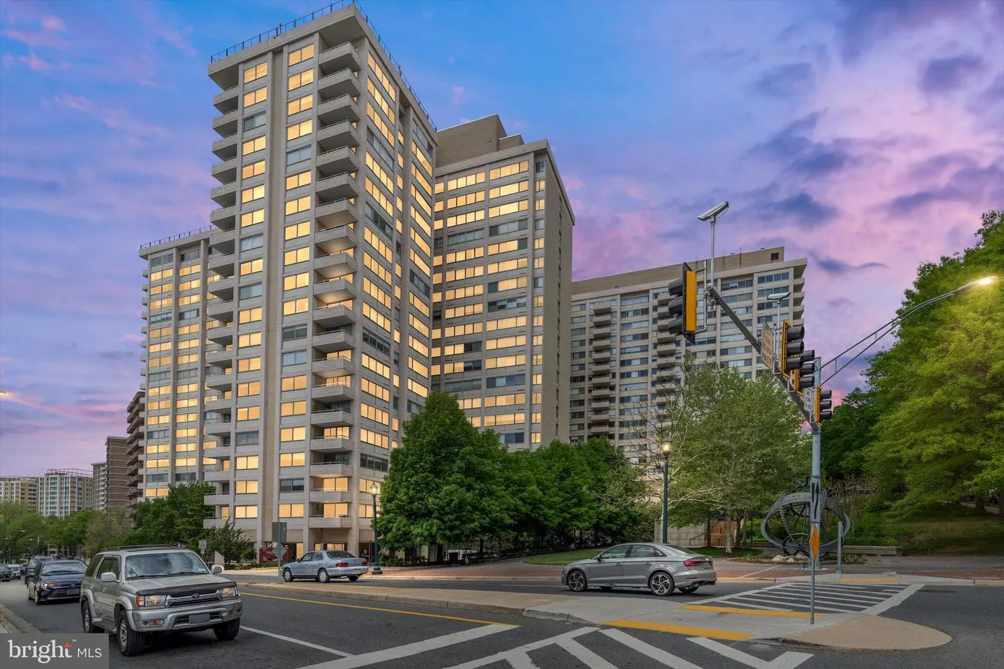 Exterior view of a tall, modern apartment building with many windows, cars on the street, and a colorful sunset sky.