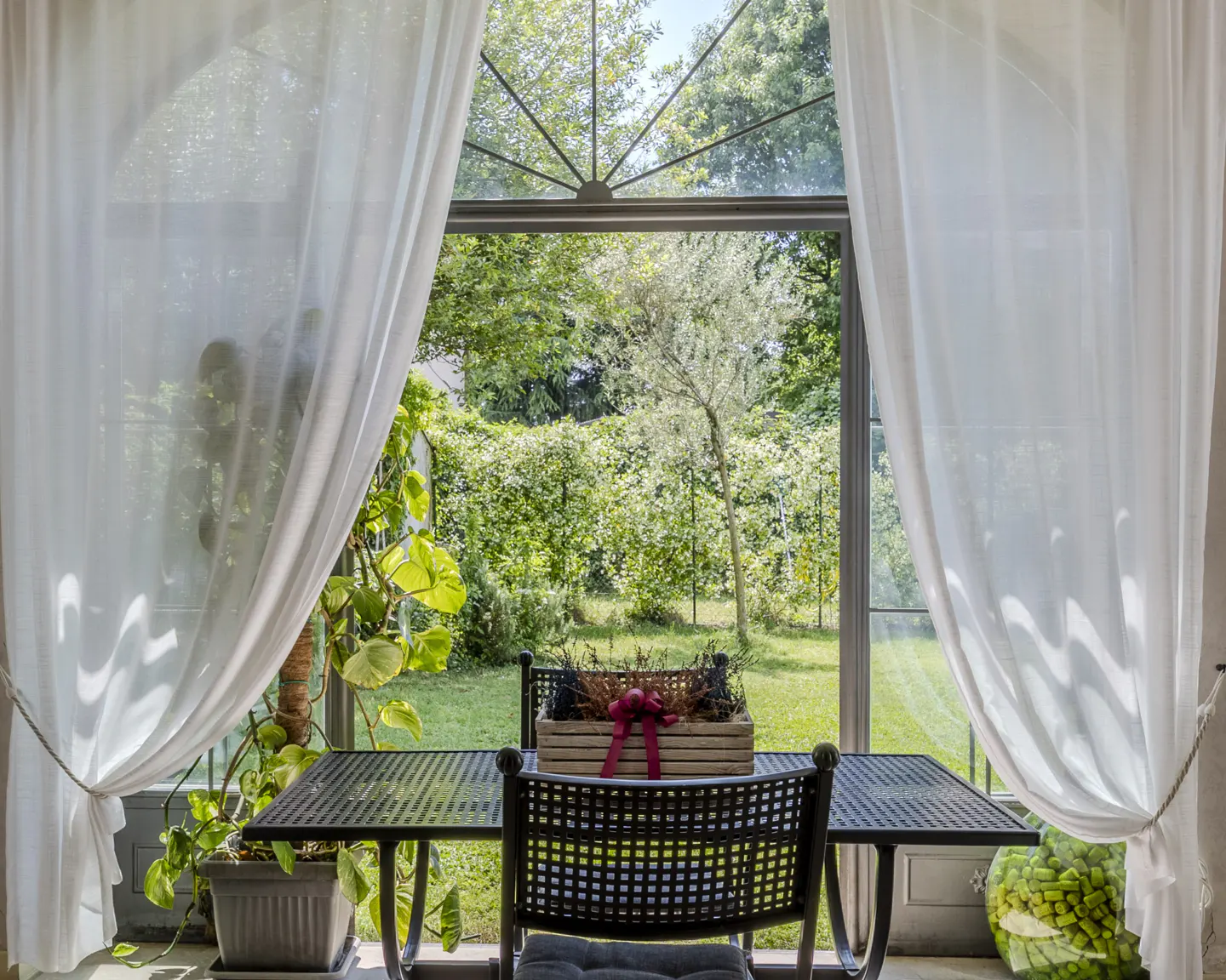 View through a window with white curtains showing a black metal table and chair overlooking a lush green garden.