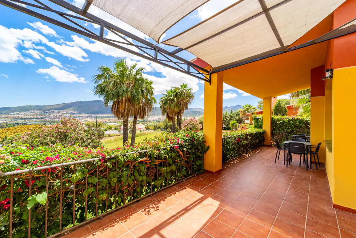 Covered patio with orange walls, brown tile floor, and metal railing with flowers. Palm trees and a distant city are visible.