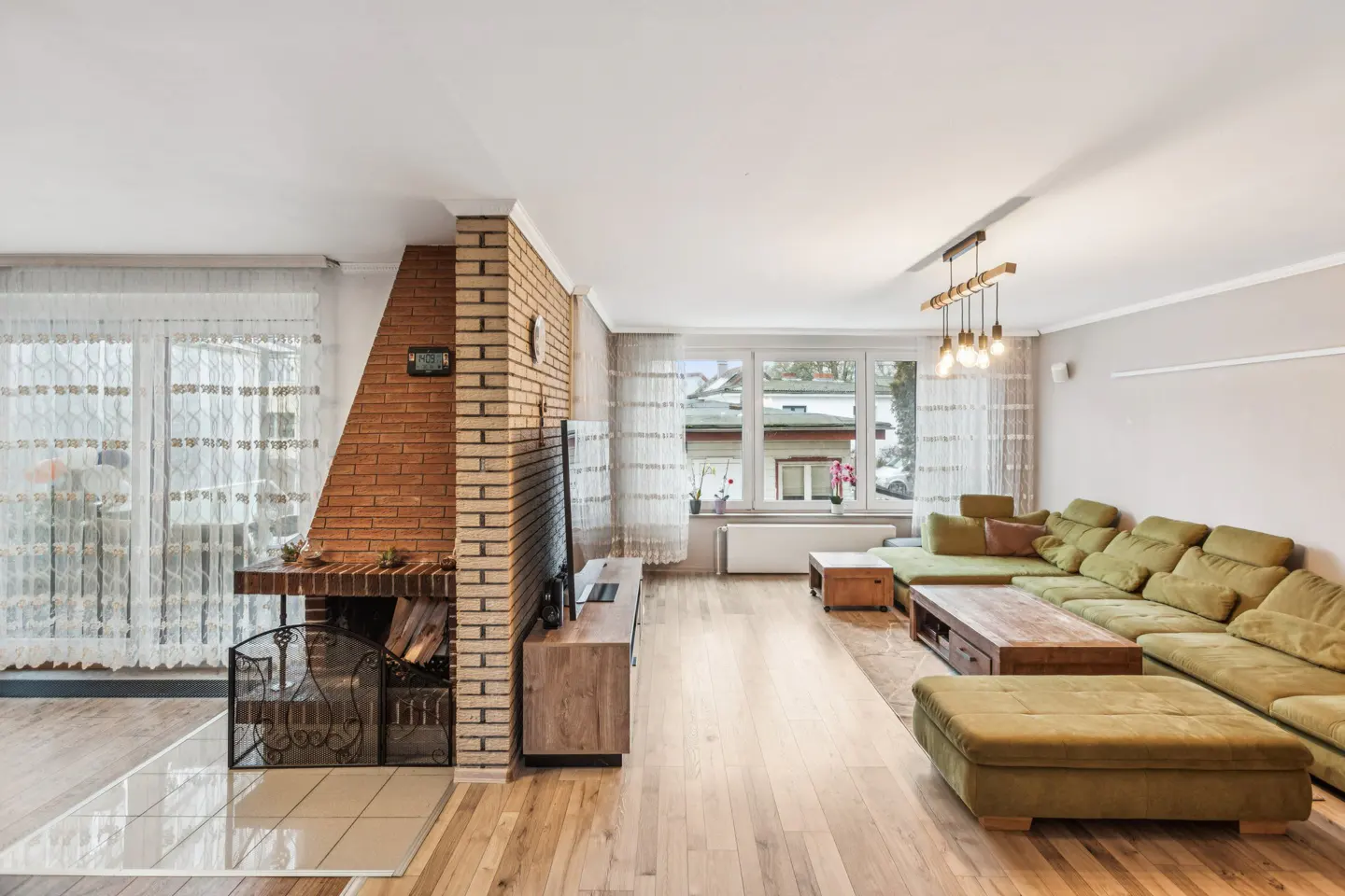 Living room with brick fireplace, hardwood floors, and a large green sectional sofa. Windows with sheer curtains let in natural light.
