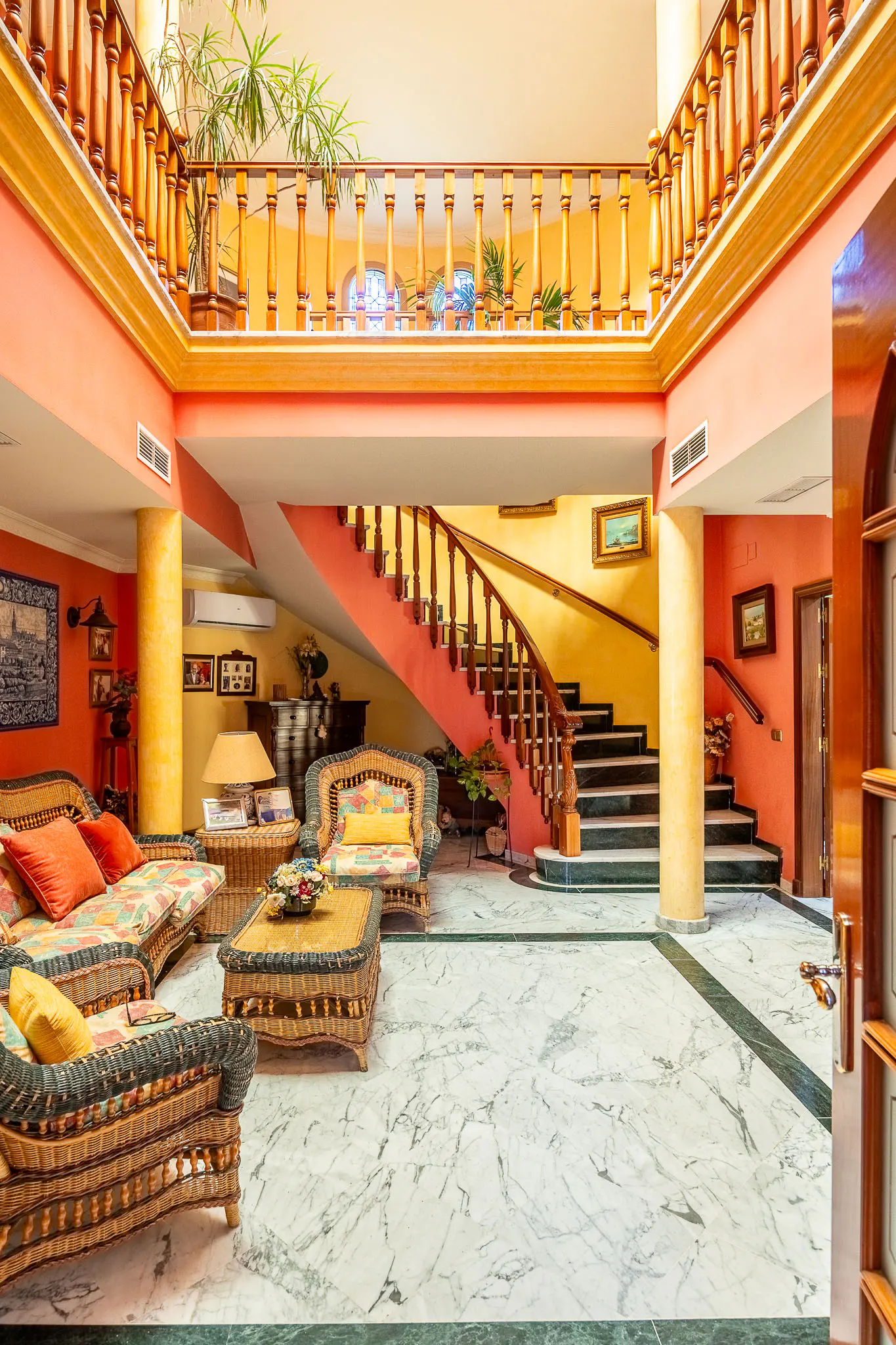 Interior view of a two-story foyer with marble floors, wicker furniture, and a curved staircase with wooden railings. Walls are painted in warm yellow and peach tones.