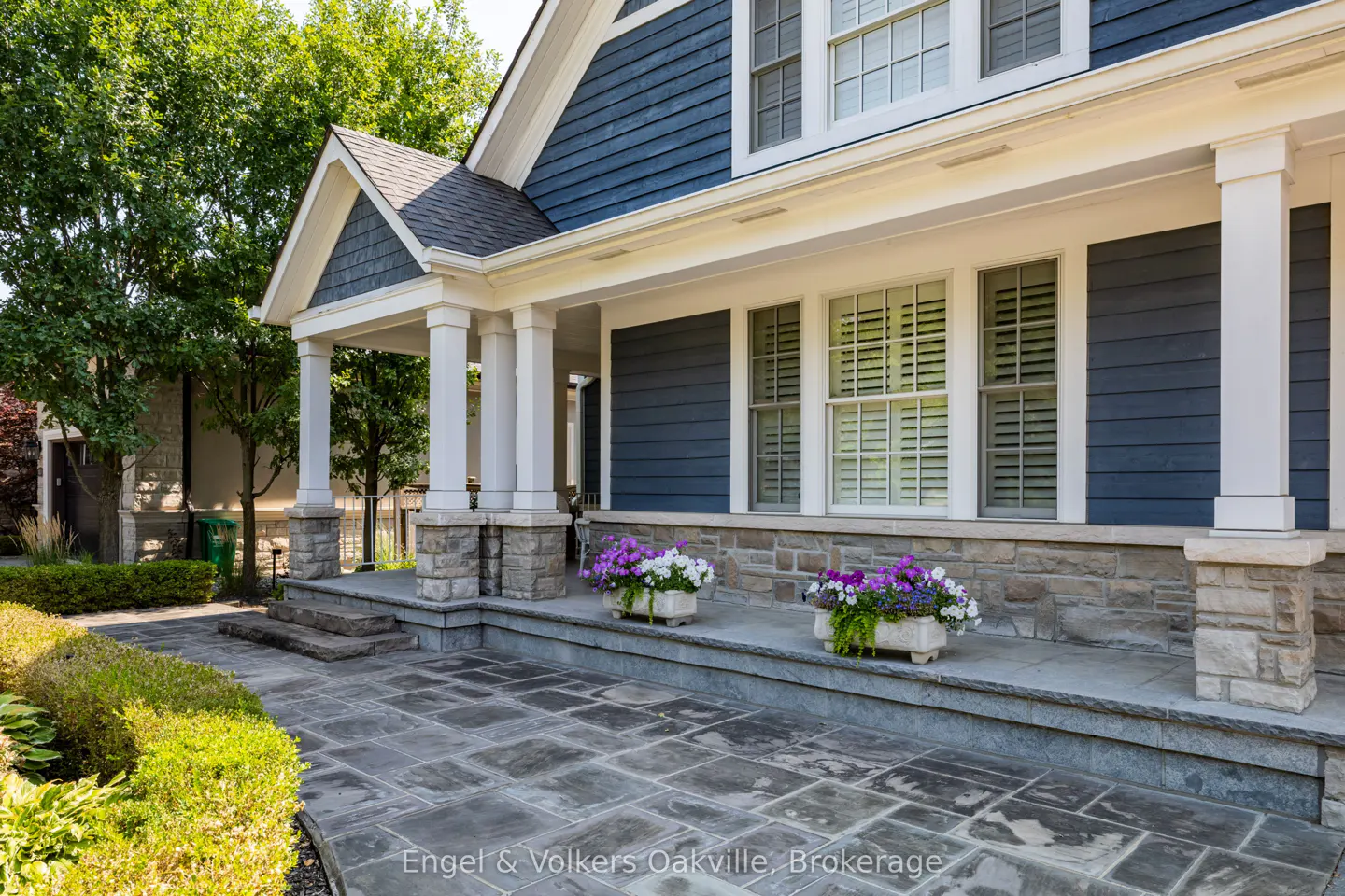 Exterior of a blue house with white trim, stone accents, and a gray stone walkway. Two flower boxes with purple and white flowers sit on the porch.